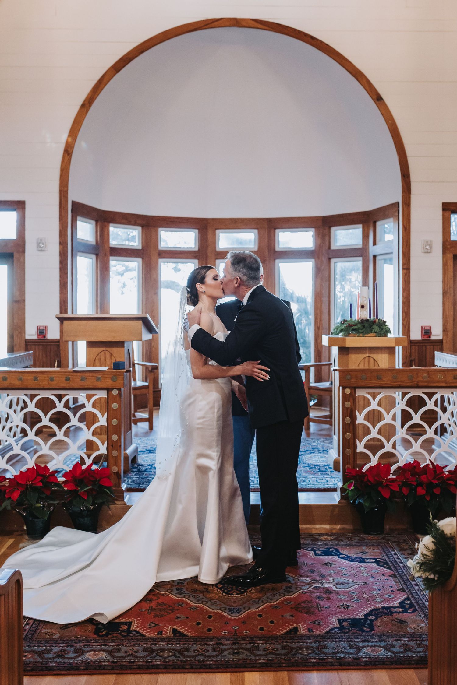 Newlyweds share first kiss at chapel altar surrounded by red poinsettias and candlelit holiday decor.