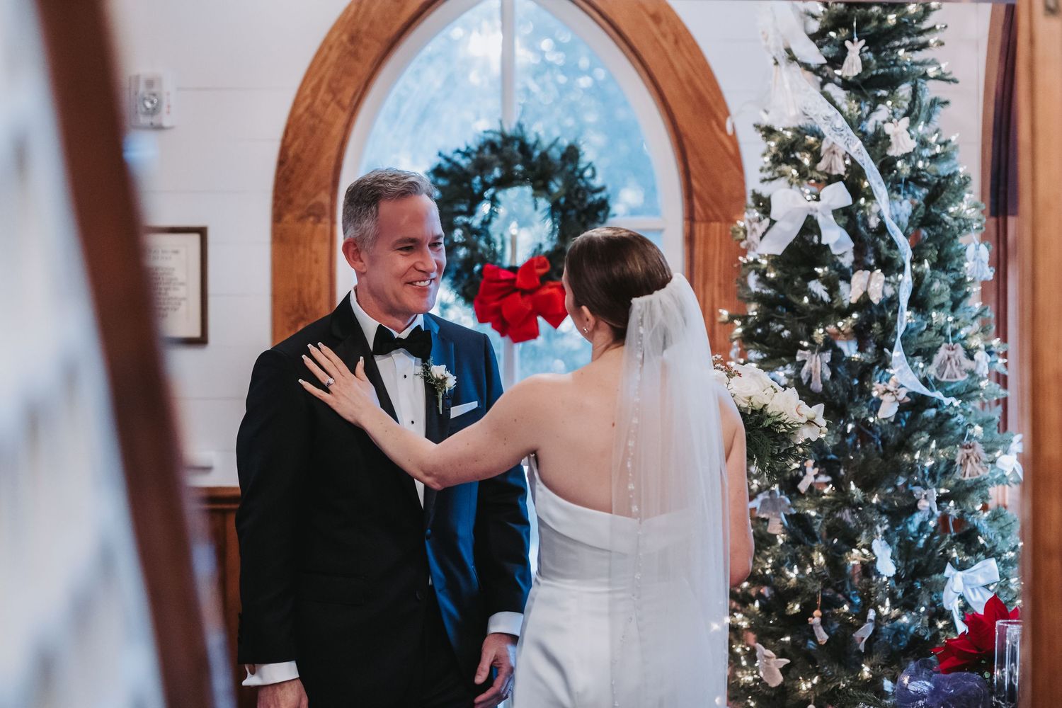 A groom smiles at his bride near a Christmas tree and wreath-adorned arched window.