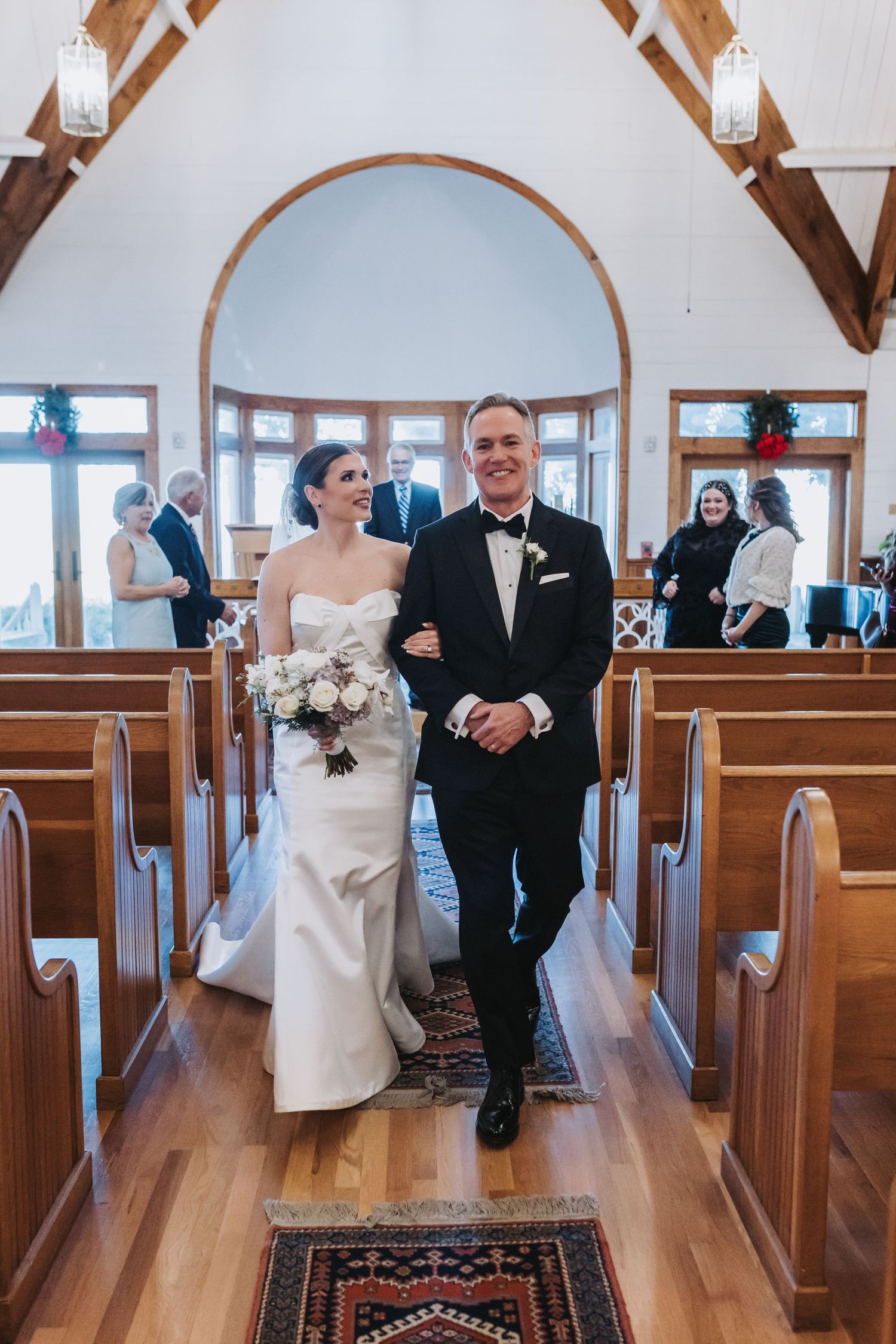 Newlyweds walking down church aisle after ceremony, bride in white gown holding bouquet, groom in tuxedo.