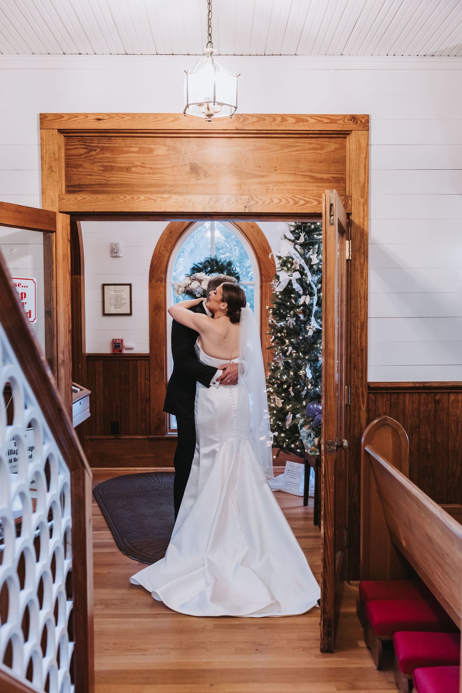 Bride and groom embrace in a charming wooden chapel doorway with a decorated Christmas tree in the background.