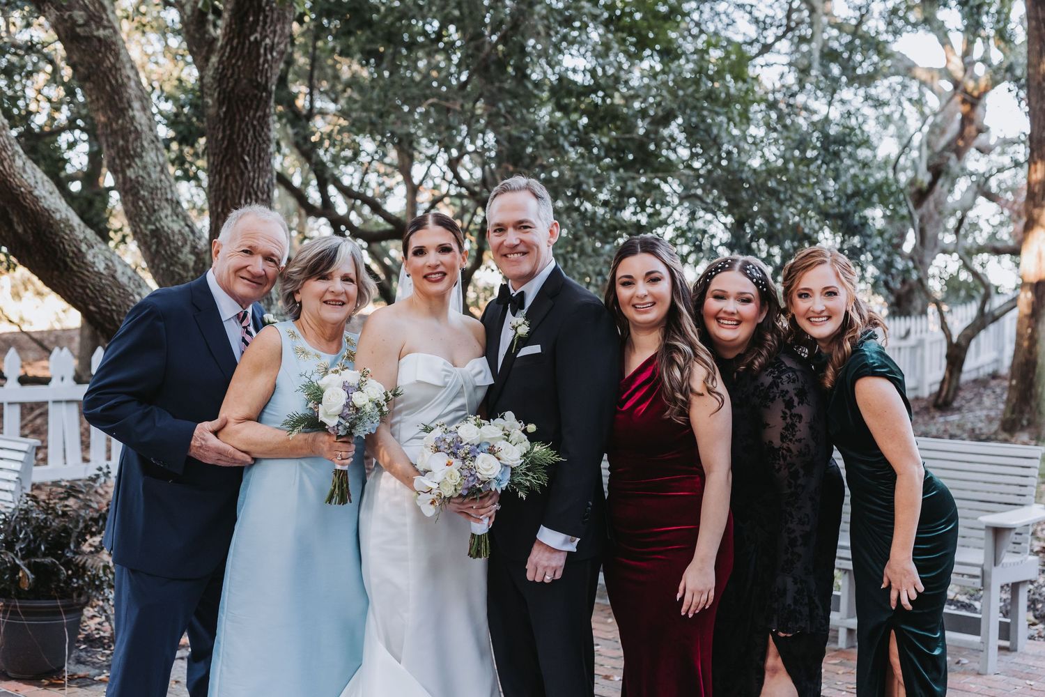 A wedding party of six poses outdoors under oak trees, bride holding a white bouquet.