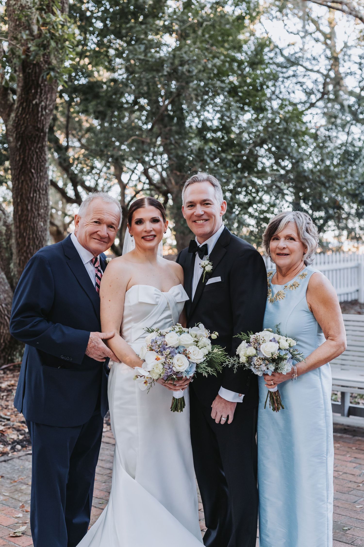 Wedding family portrait with bride in white gown and groom in tuxedo with parents outdoors.
