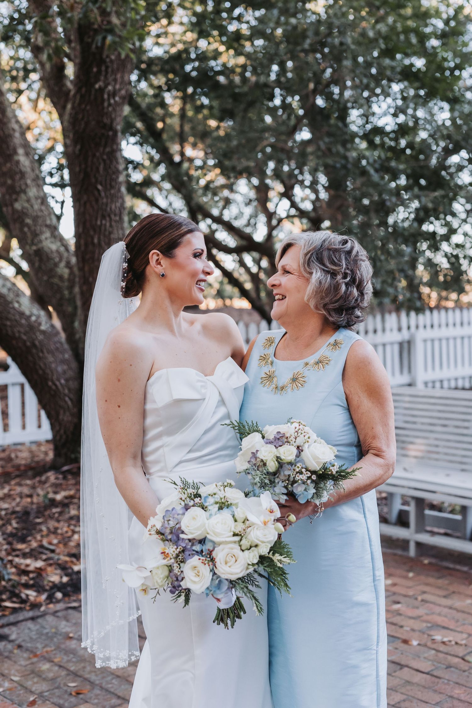 Bride in white strapless gown and veil with older woman in light blue dress holding bouquets.