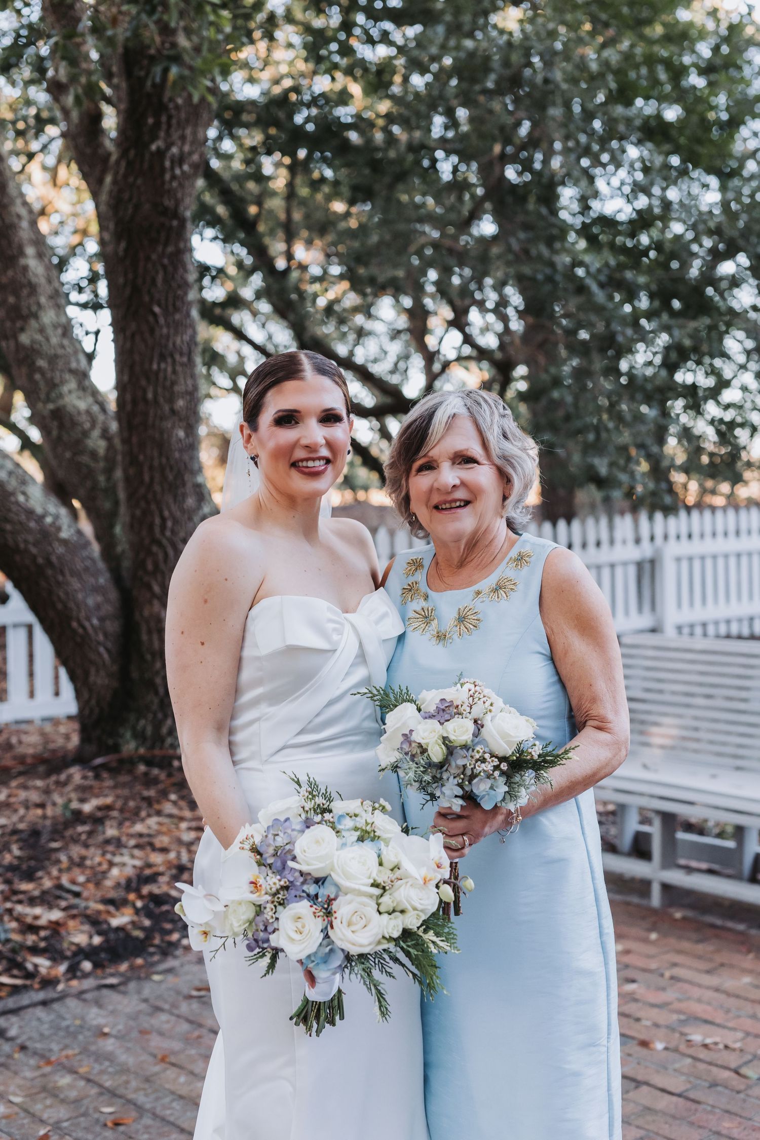 Bride in strapless white gown and older woman in light blue dress holding white floral bouquets outdoors.