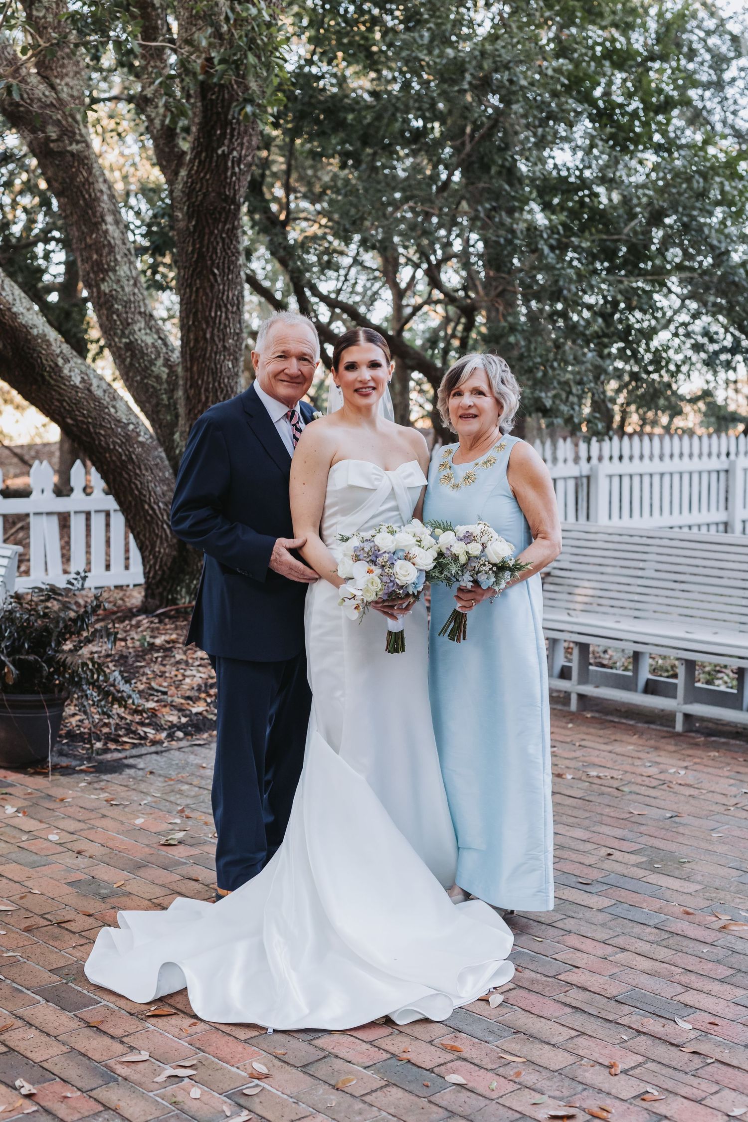 Bride in strapless white gown poses with parents outdoors on brick patio beneath oak trees.