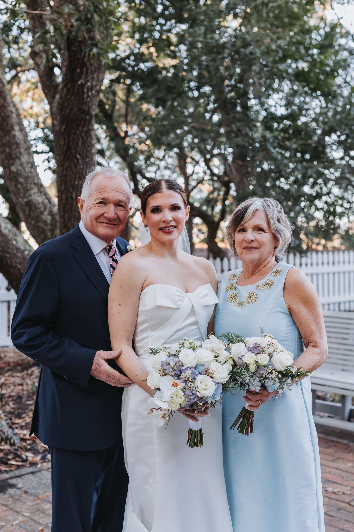 Bride in strapless white gown poses with parents outdoors, mother holding white floral bouquet in light blue dress.