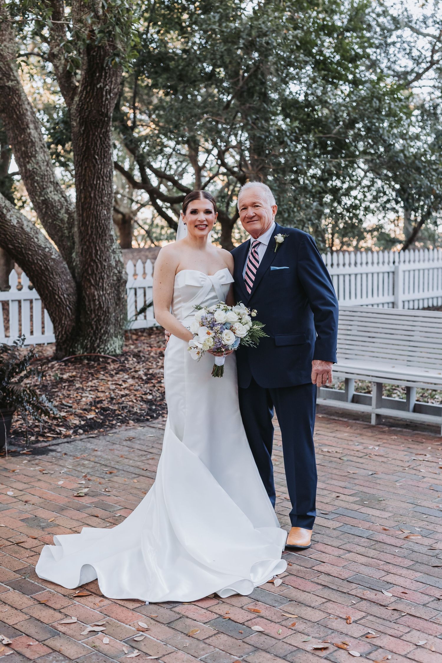Bride in elegant white mermaid gown poses with father in navy suit on brick pathway under oak trees.