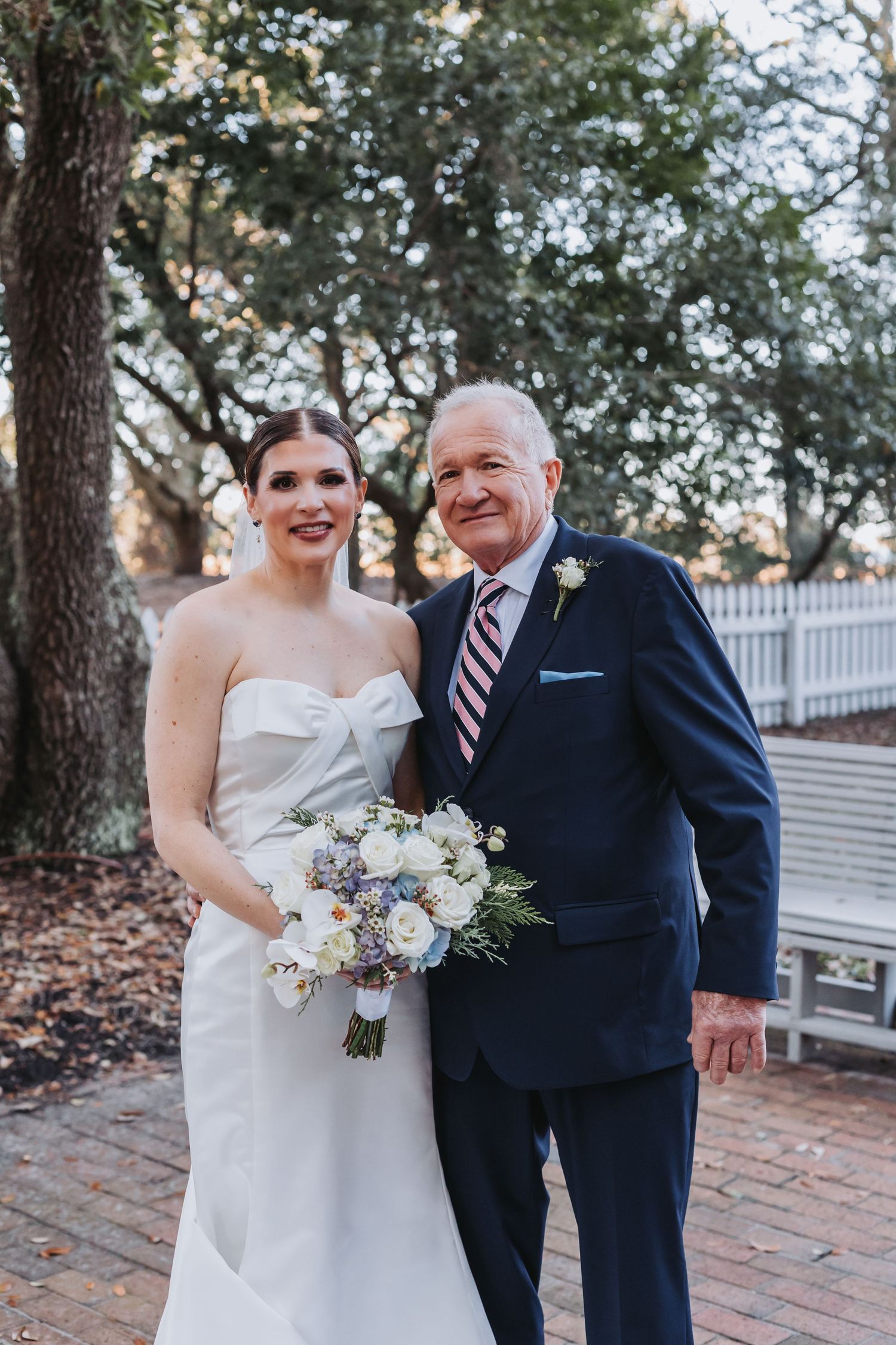 Smiling bride in strapless white gown poses with older gentleman in navy suit holding bouquet outdoors.