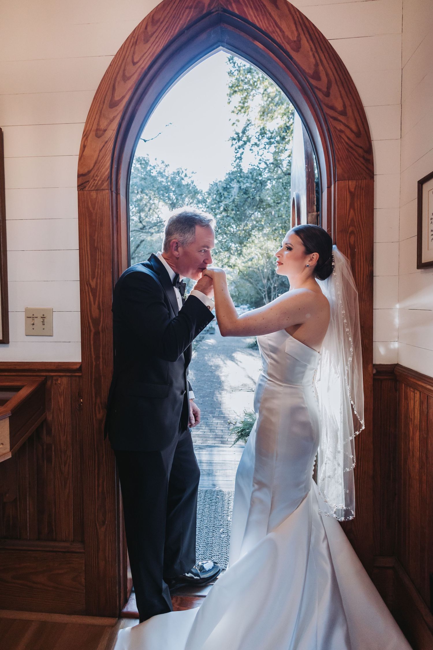 Groom in tuxedo kisses bride's hand in romantic moment framed by arched wooden church window.