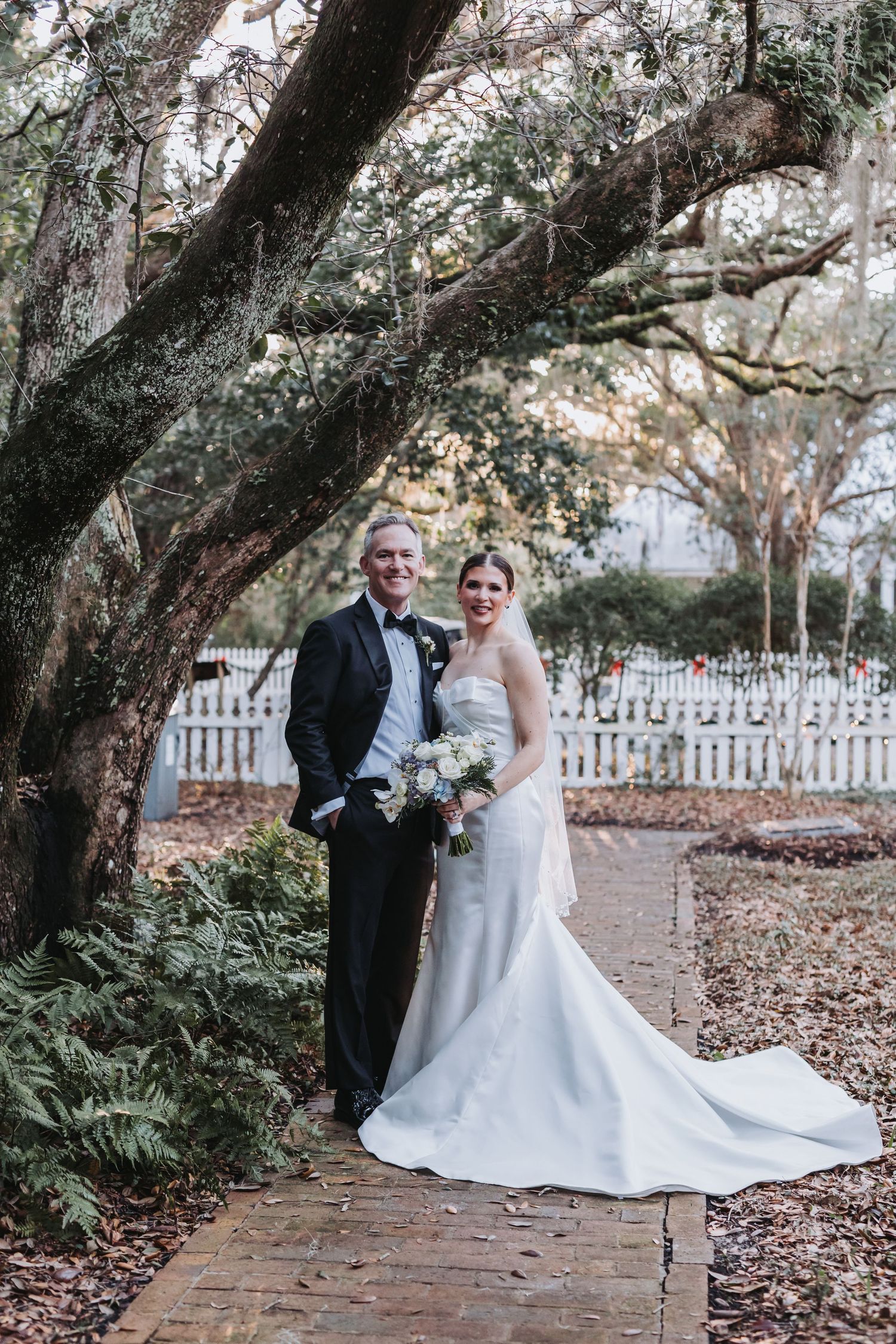 Bride and groom posing under moss-covered oak tree on brick path with white picket fence backdrop.