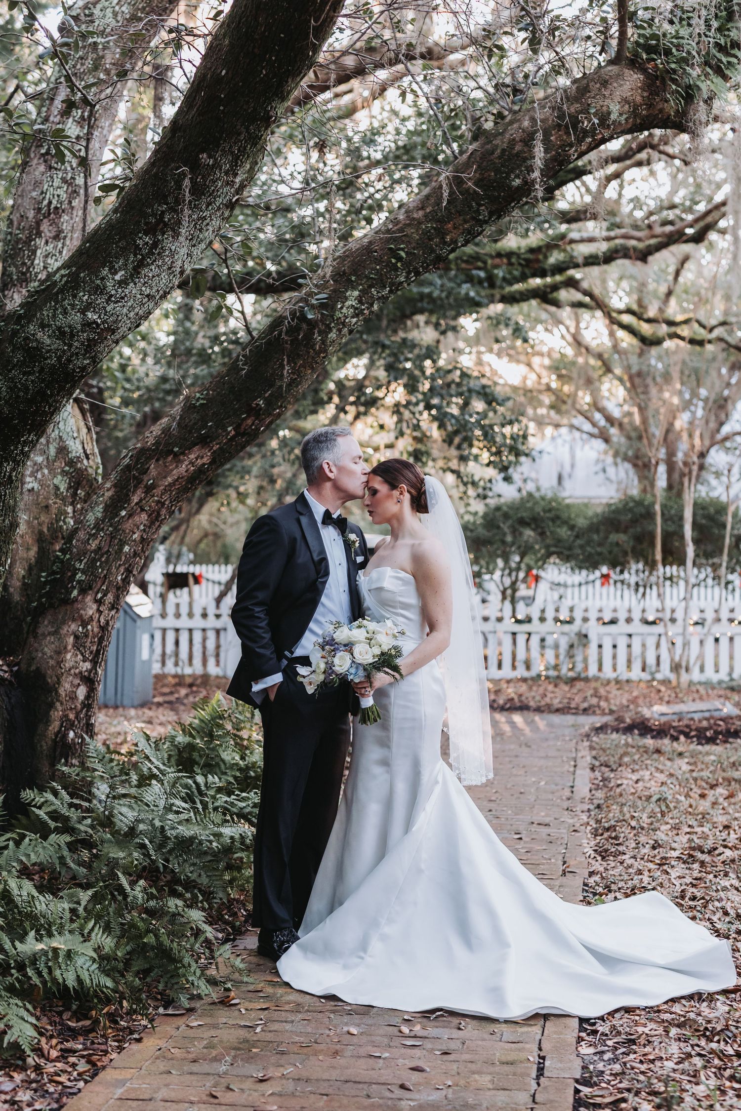 Bride and groom kiss under sprawling oak tree draped with Spanish moss on brick garden path.