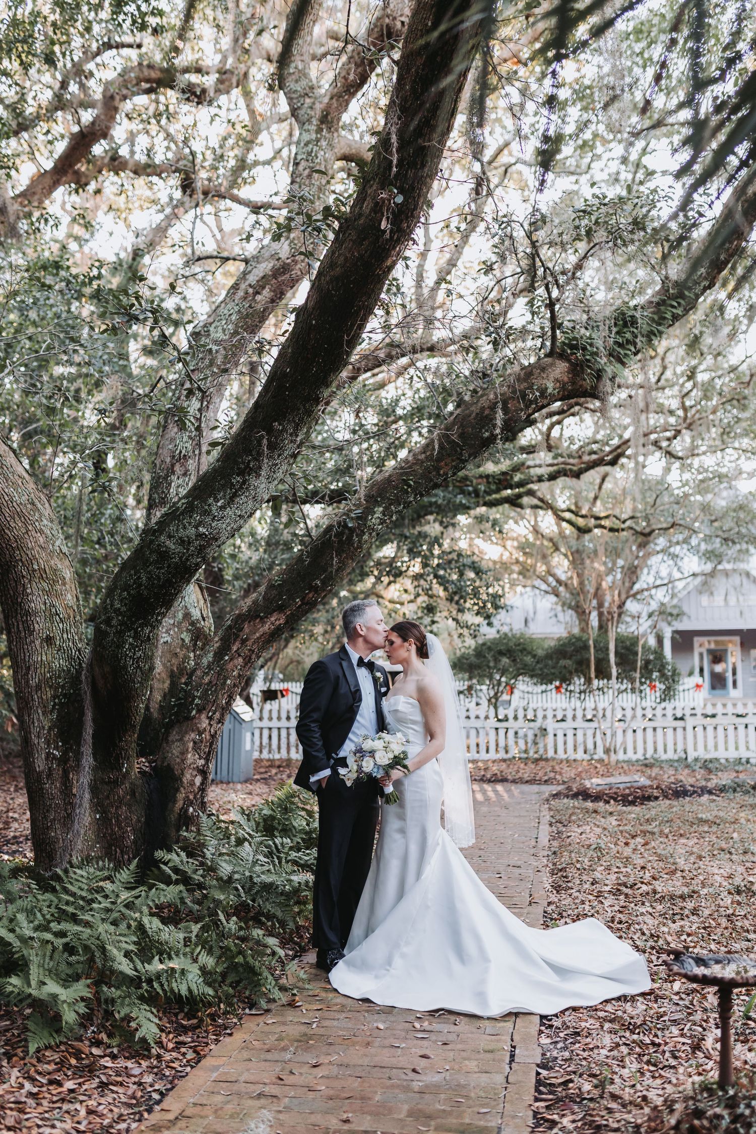 Newlyweds embrace and kiss beneath large oak tree with Spanish moss on rustic brick pathway.