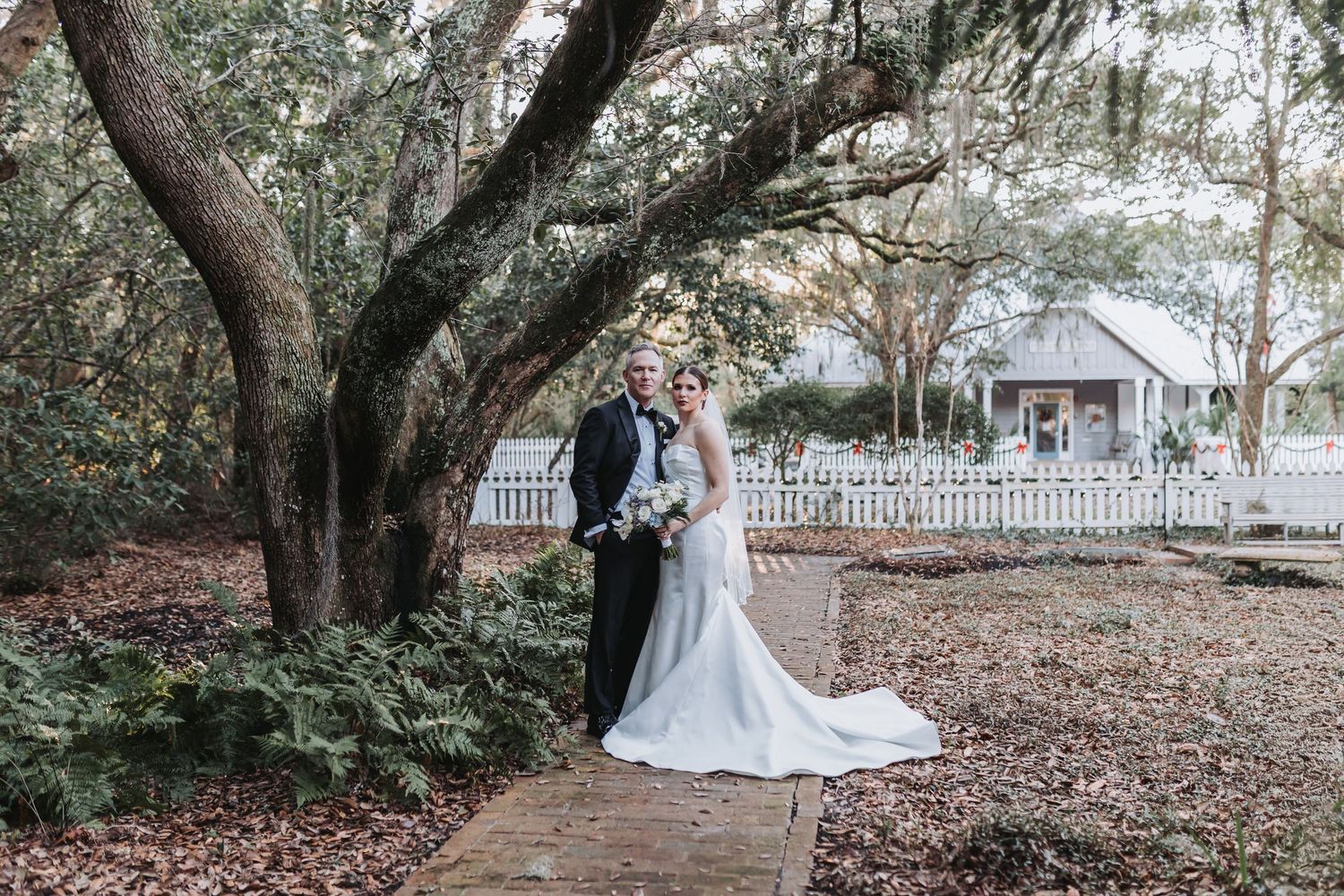A bride and groom pose together on a garden path beneath a large oak tree with a white fence backdrop.