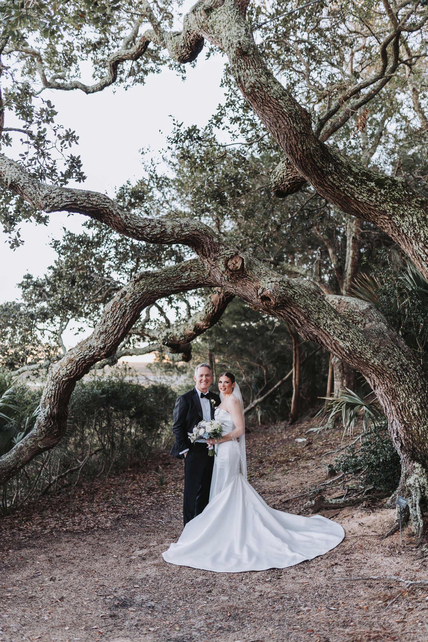 Bride and groom pose under sprawling oak tree in natural outdoor wedding portrait.