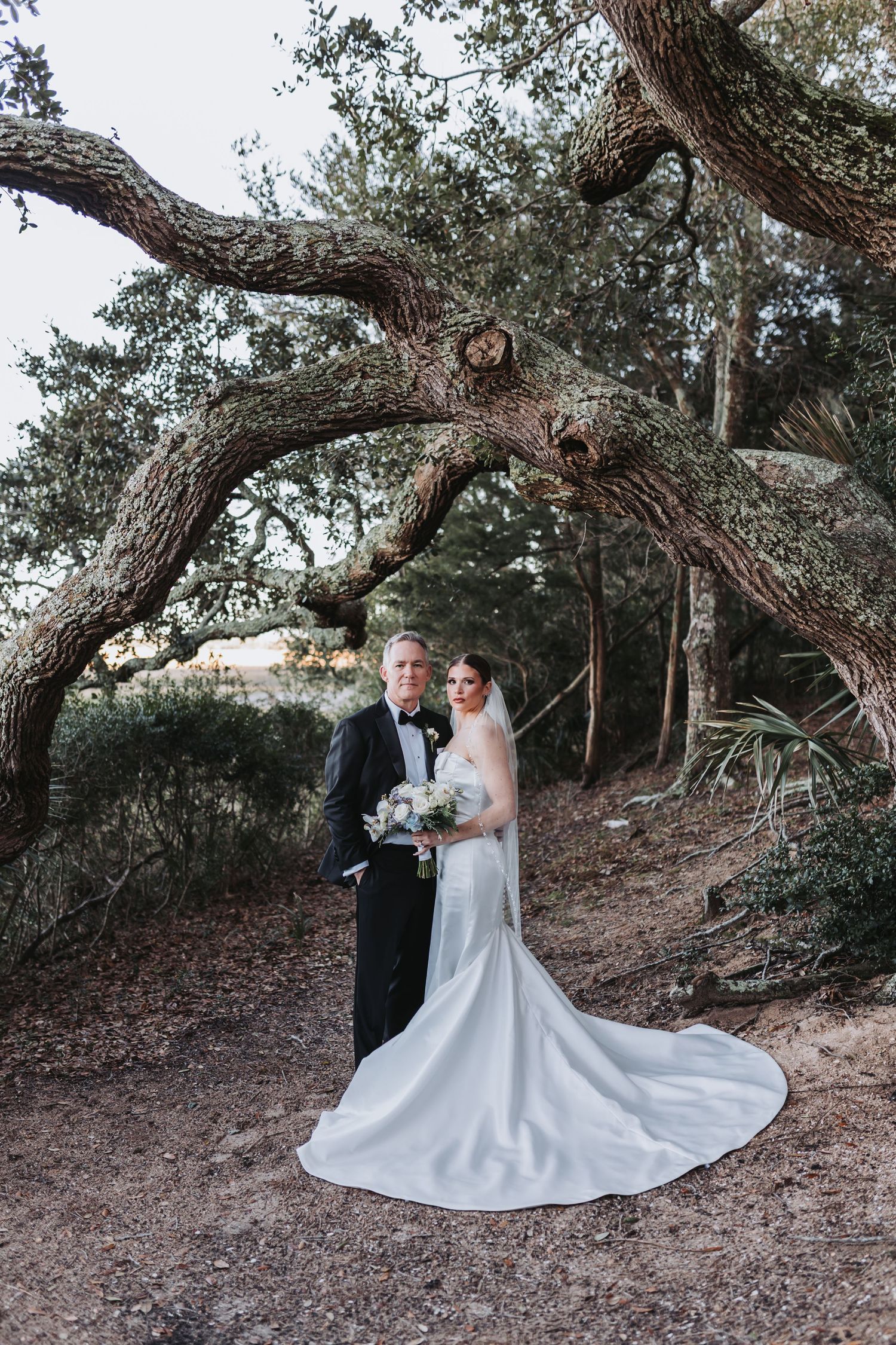 Elegant bride and groom stand beneath twisted oak tree with flowing wedding dress train.