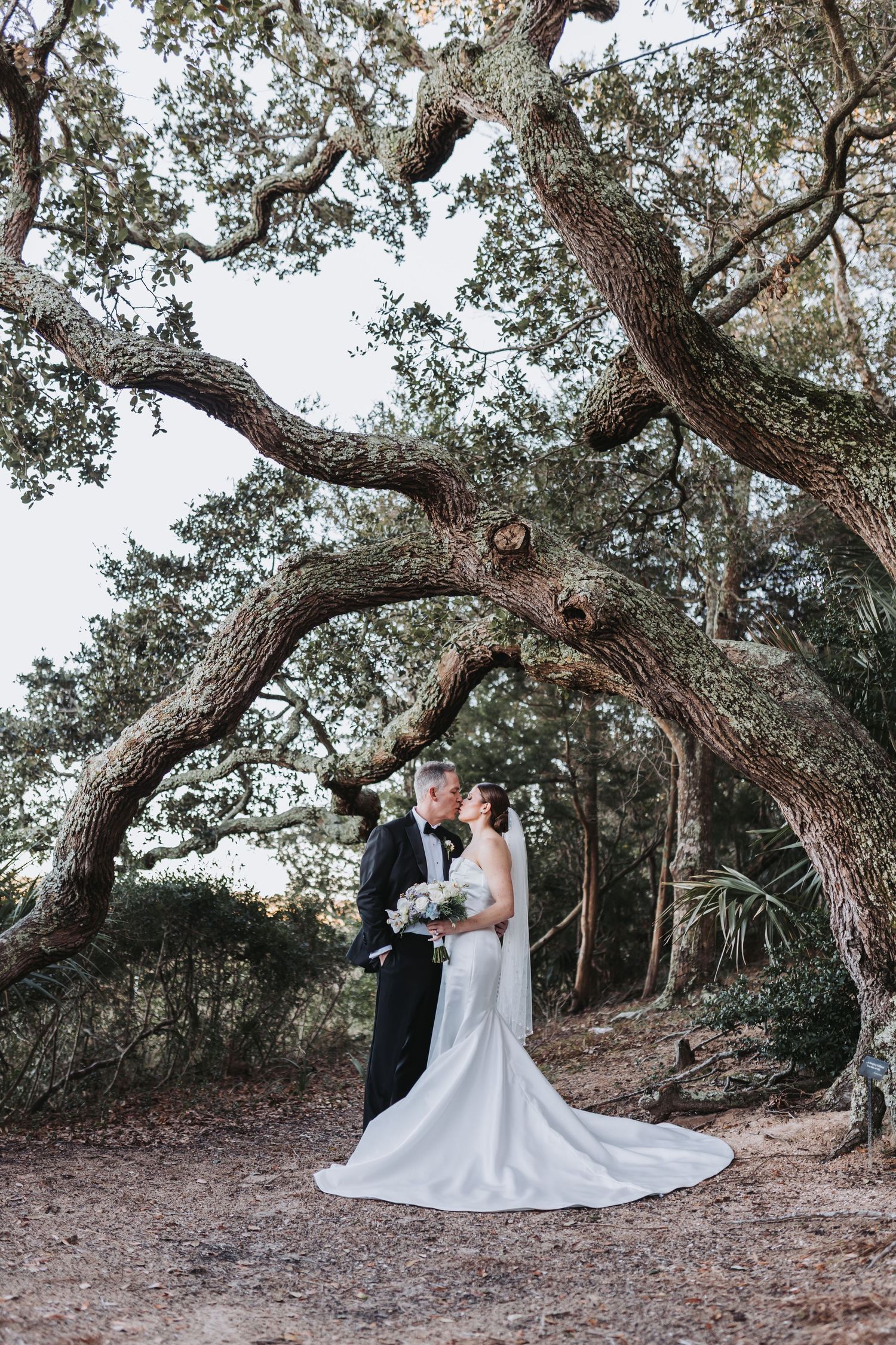 Bride and groom share a romantic kiss beneath a sprawling oak tree in a lush outdoor wedding portrait.