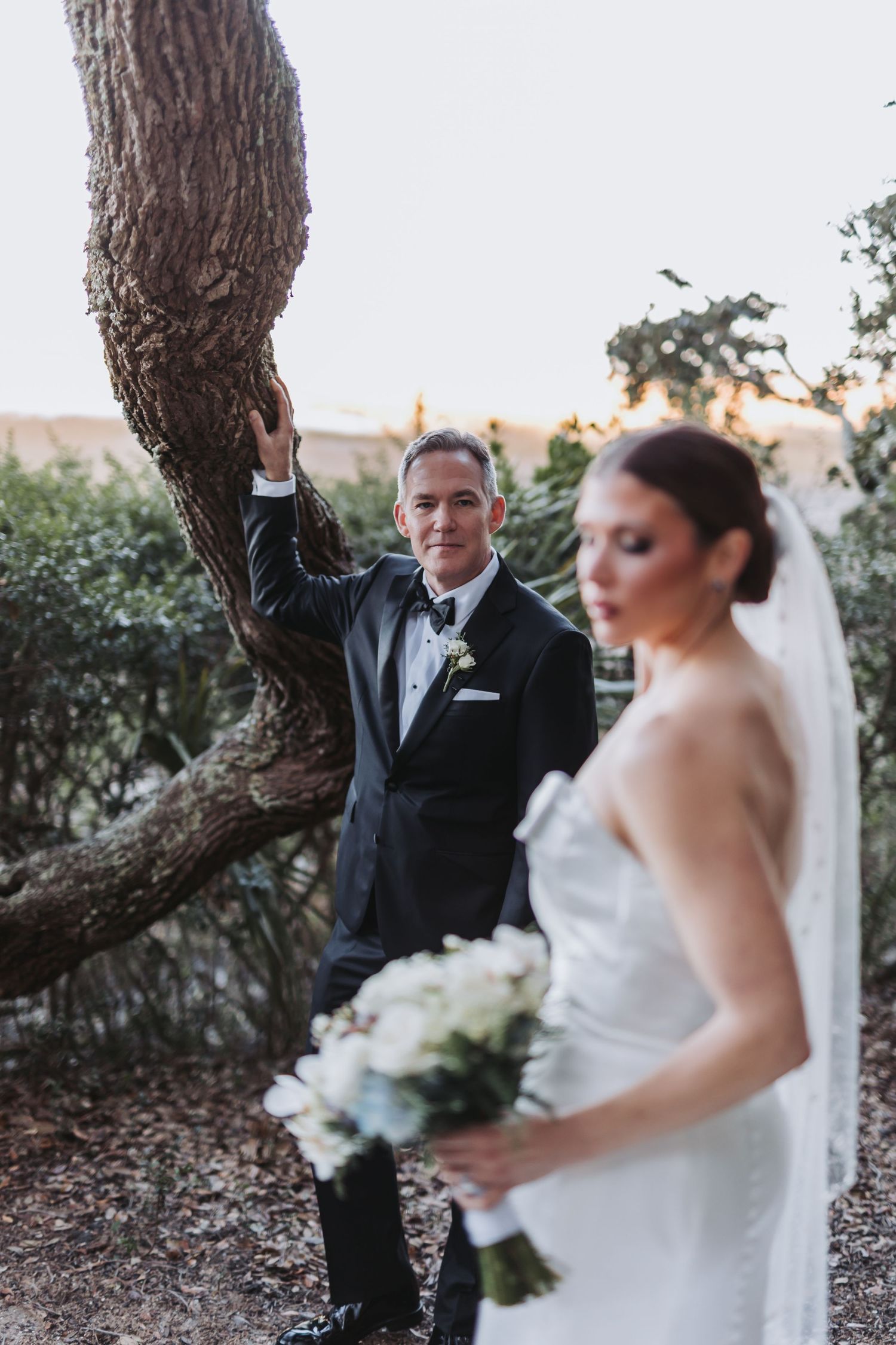 Groom in black tuxedo leans against oak tree while bride holds white bouquet in foreground.