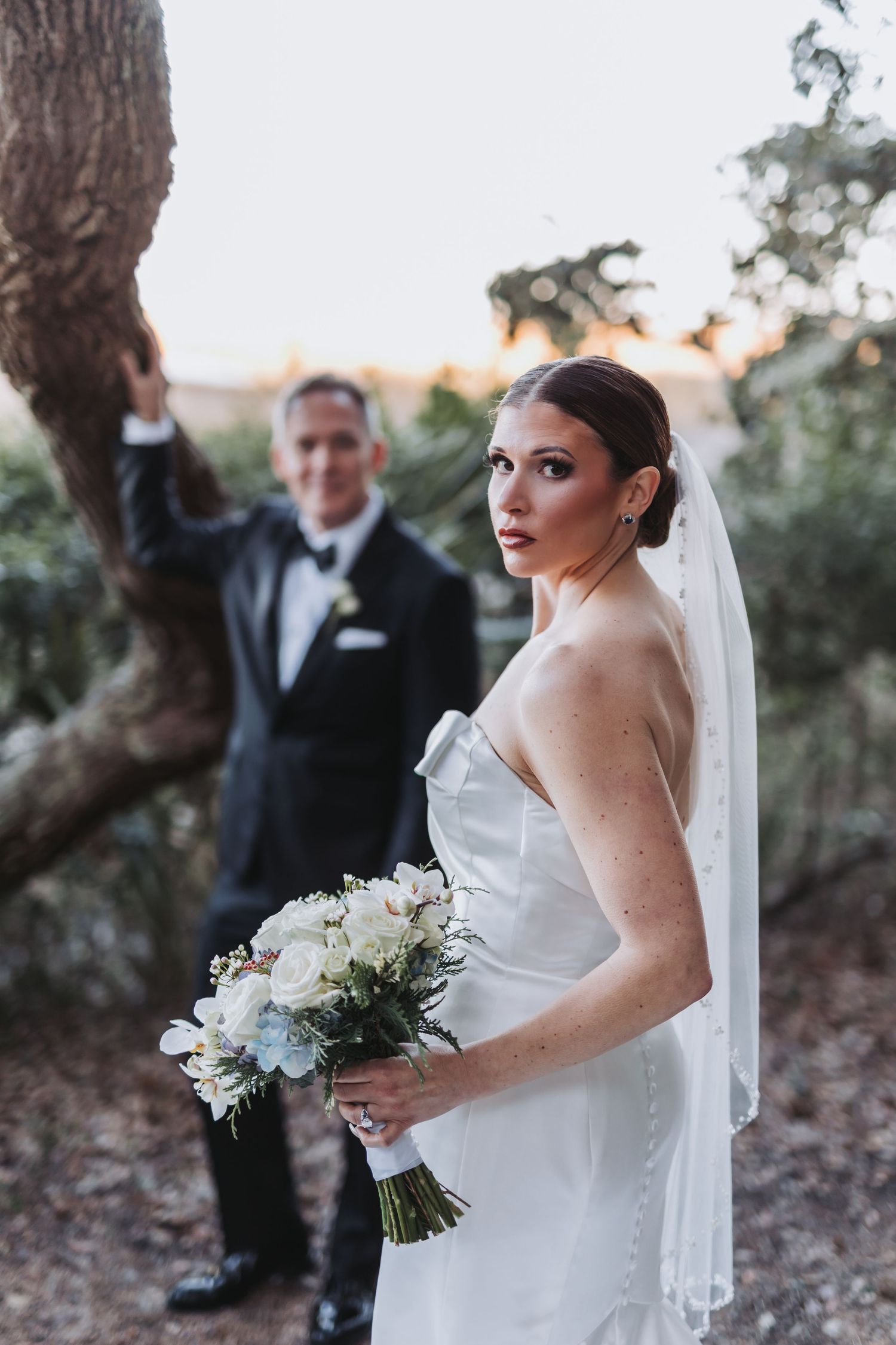 Elegant bride in a strapless gown holding a bouquet while groom stands beneath a tree at sunset.