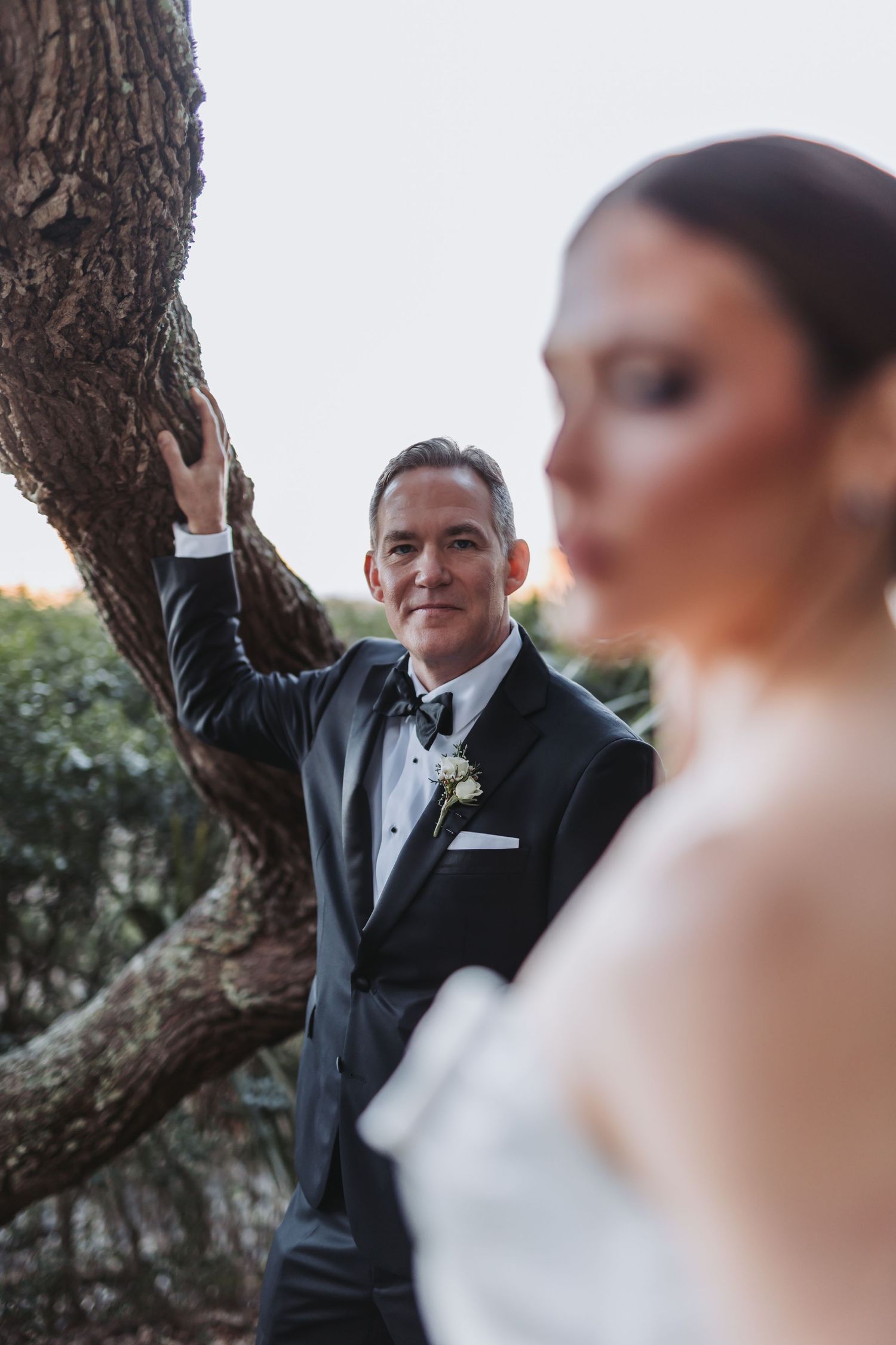 Groom in black tuxedo leaning against oak tree gazing at bride in white dress in foreground.