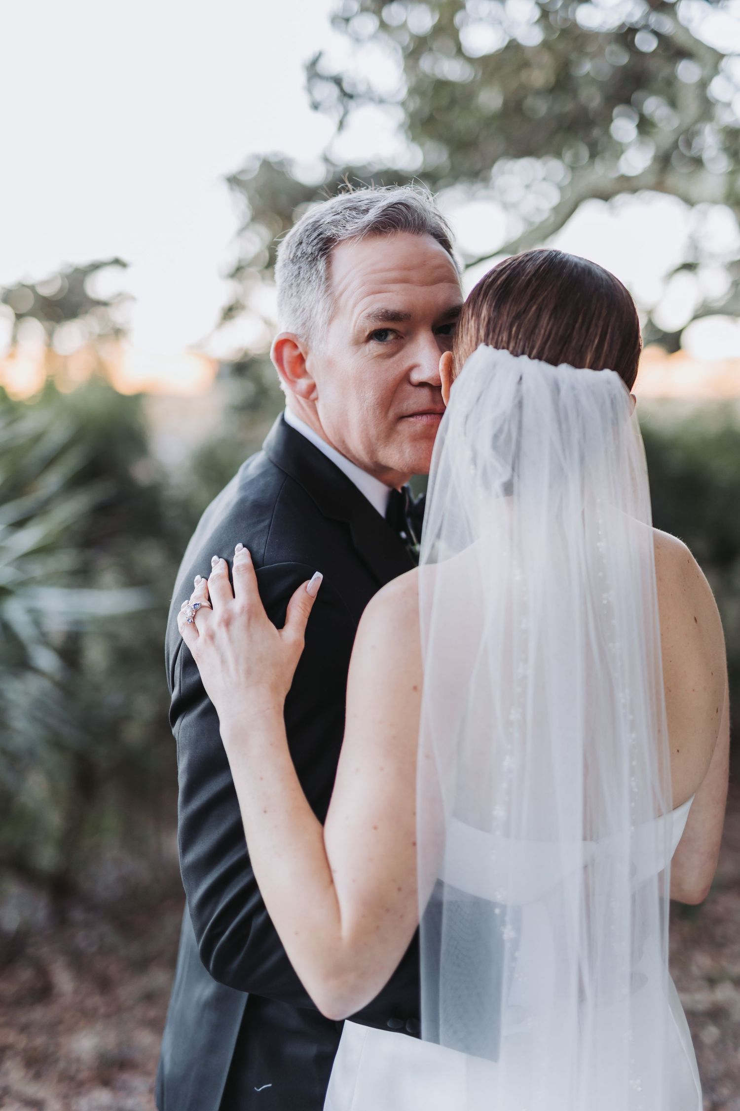 Emotional father embraces bride in veil during tender outdoor wedding moment at sunset.