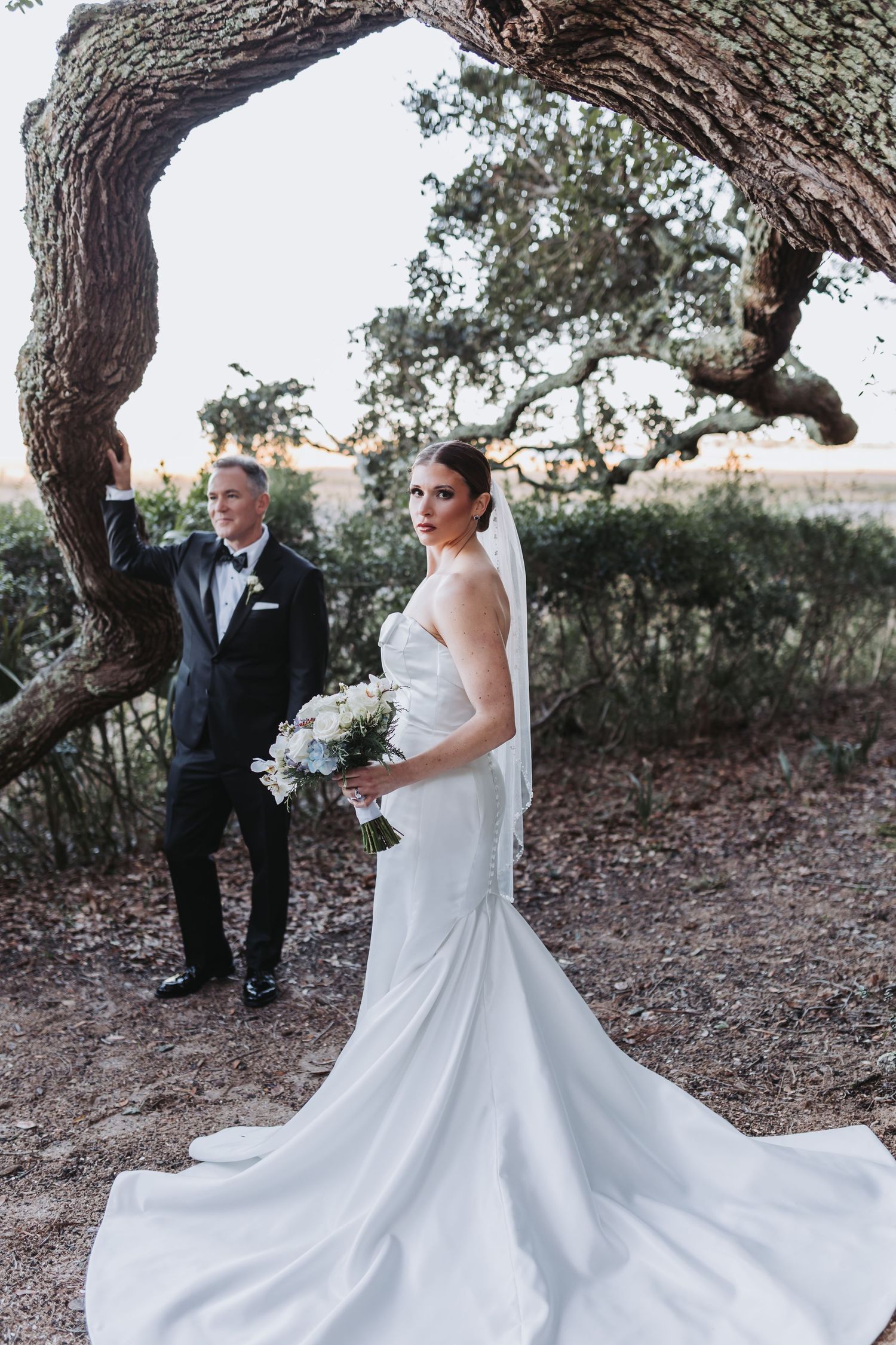 A bride in a flowing white gown holds a bouquet while her groom in a tuxedo stands beneath a twisted oak tree.