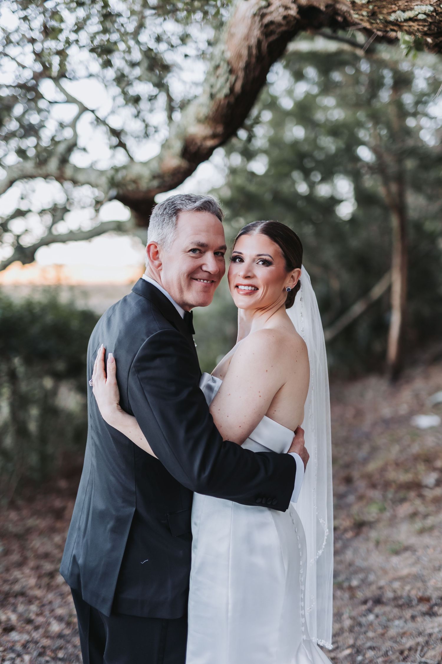 Smiling bride and groom embrace outdoors under oak tree canopy in classic black tuxedo and white gown.