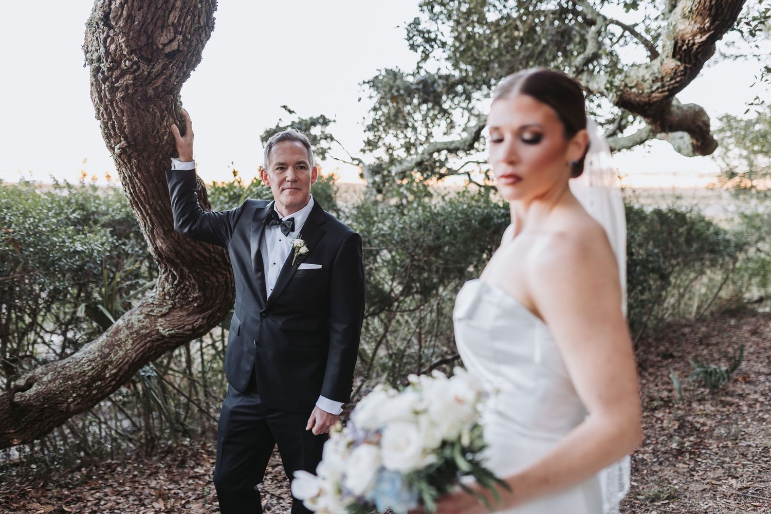 Groom in black tuxedo leans against oak tree as bride in strapless gown holds white bouquet.