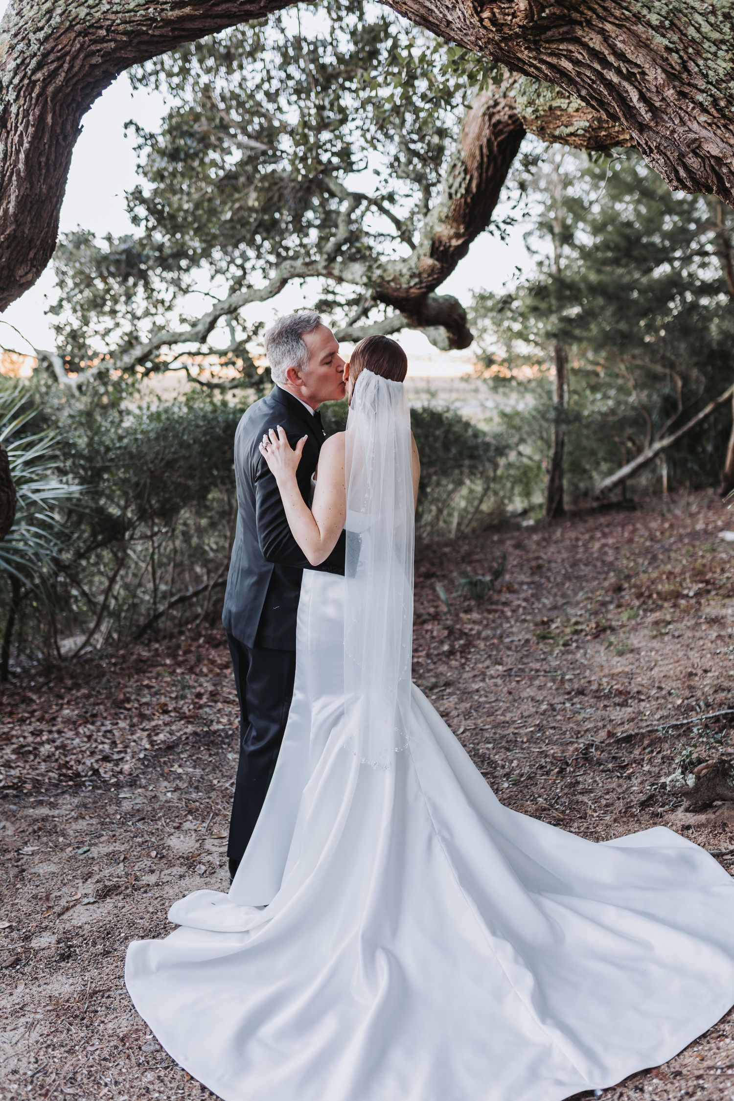 A bride and groom share a romantic kiss beneath a large oak tree with the bride's veil trailing behind.