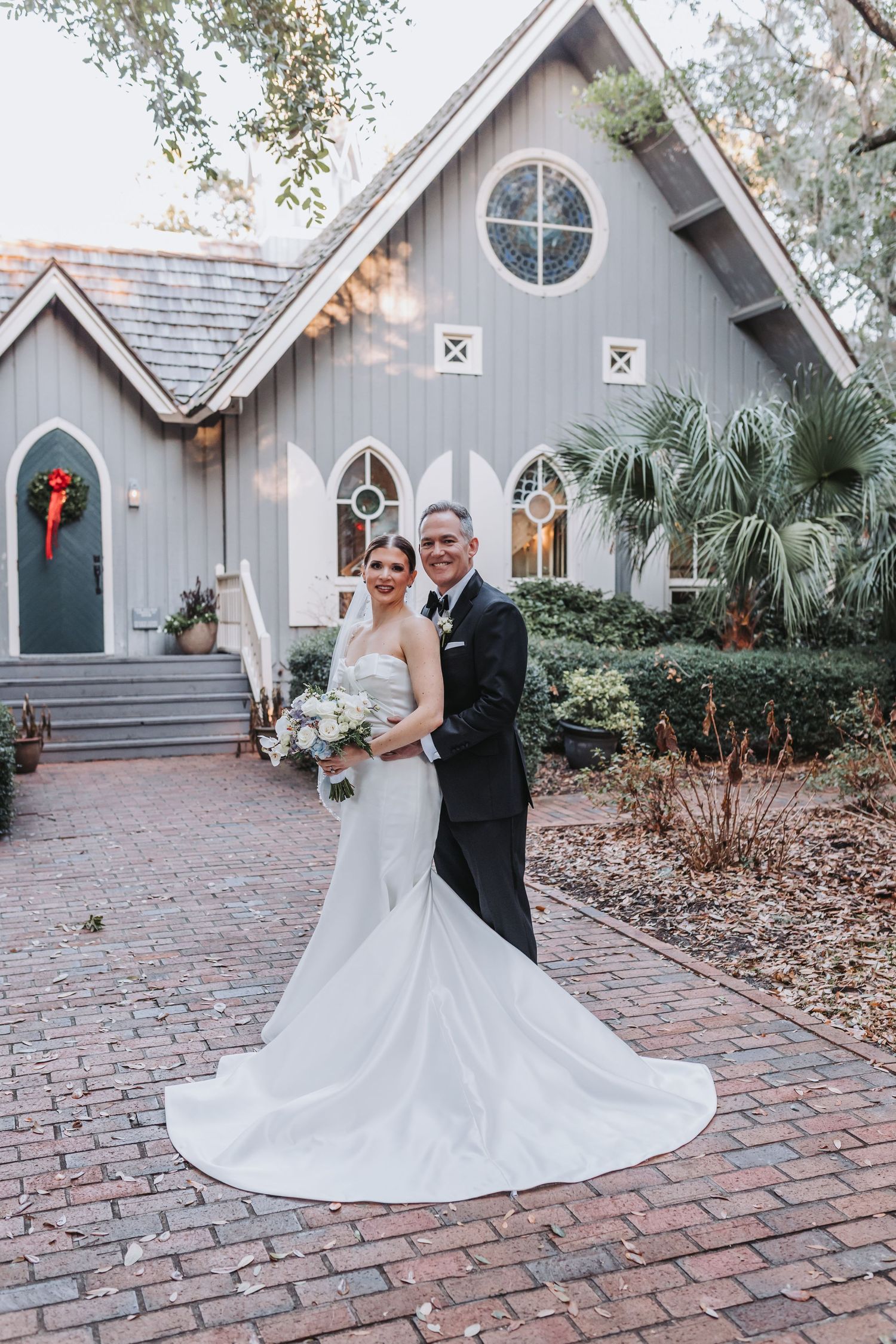 Newlyweds pose together in front of charming gray chapel with Christmas wreath on brick pathway.