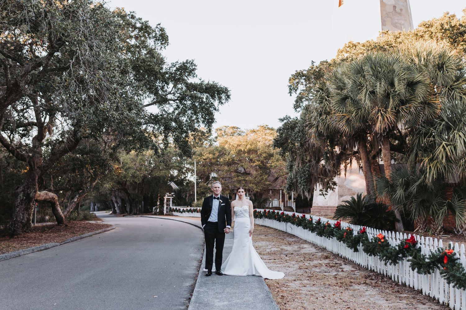 Bride and groom walking together on tree-lined road with white fence and holiday decorations.