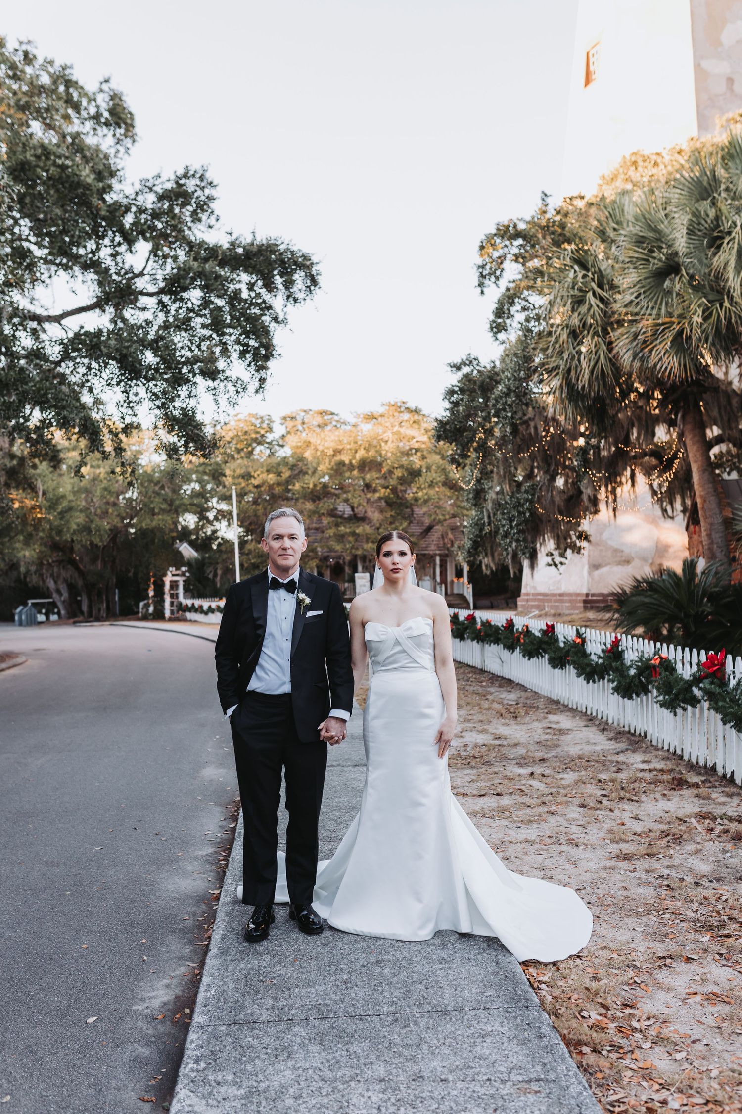 Bride and groom in formal wedding attire hold hands while standing on sidewalk lined with holiday garland.