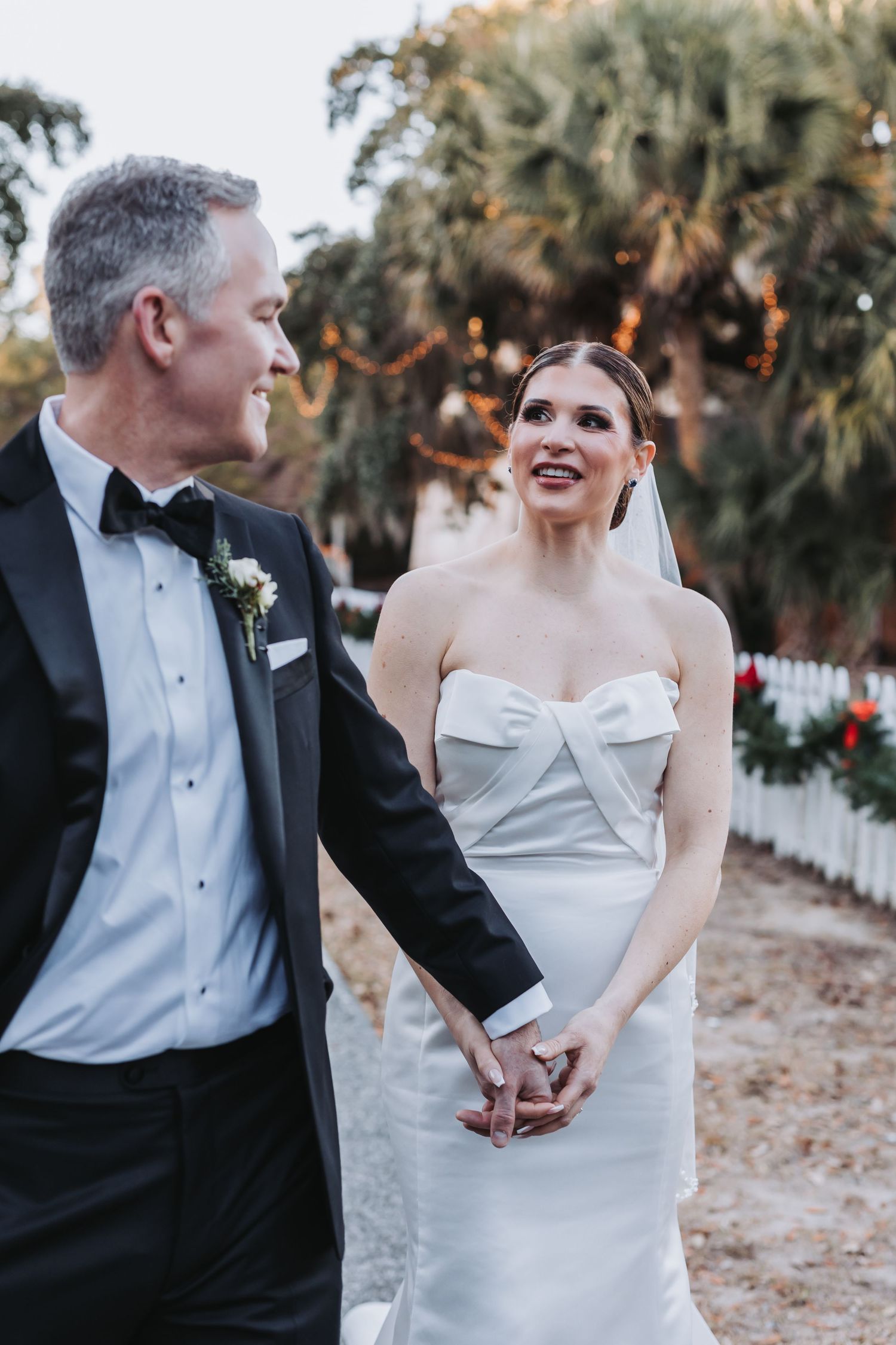 Smiling bride in strapless white gown holds grooms hand while walking outdoors at golden hour.