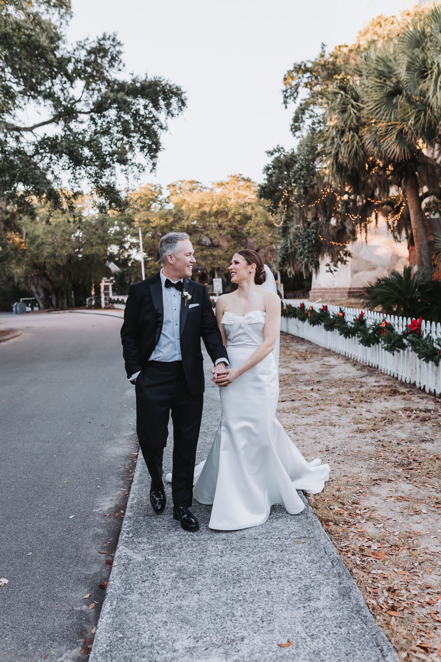 Bride and groom walk hand-in-hand along a sidewalk lined with white picket fence and holiday greenery at golden hour.