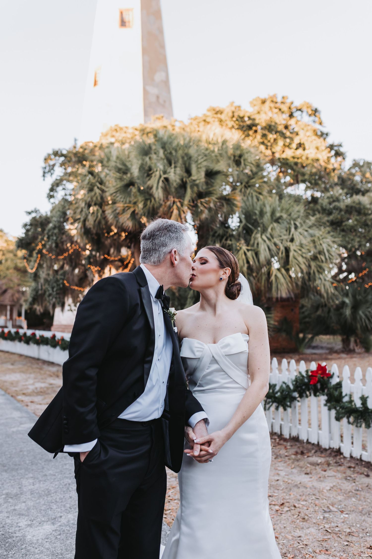 Bride and groom kissing near a lighthouse with Christmas garland on a white picket fence at golden hour.