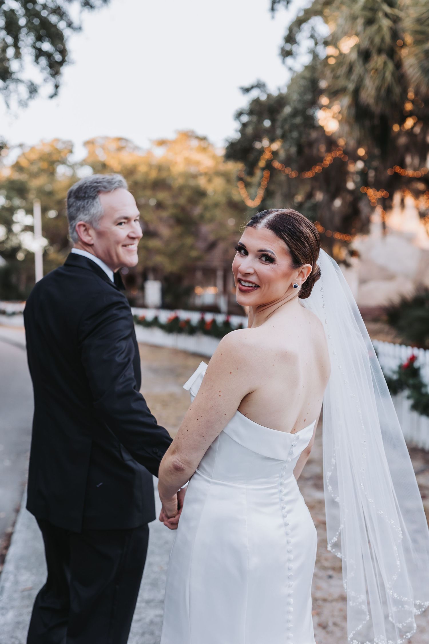 Bride in strapless white gown with veil glances back smiling while holding grooms hand outdoors.