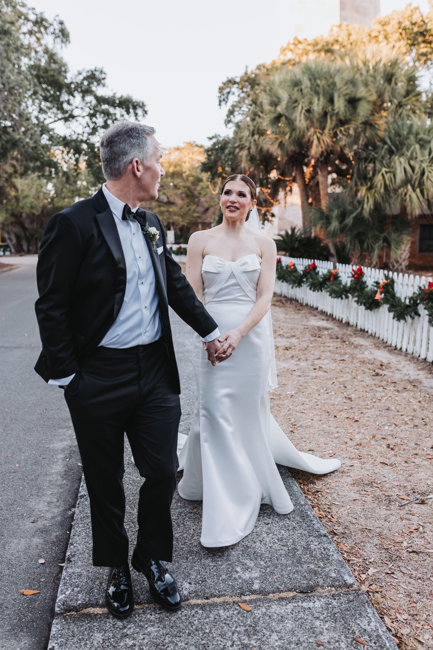 Bride and groom walk hand in hand along sidewalk lined with holiday garland at golden hour.