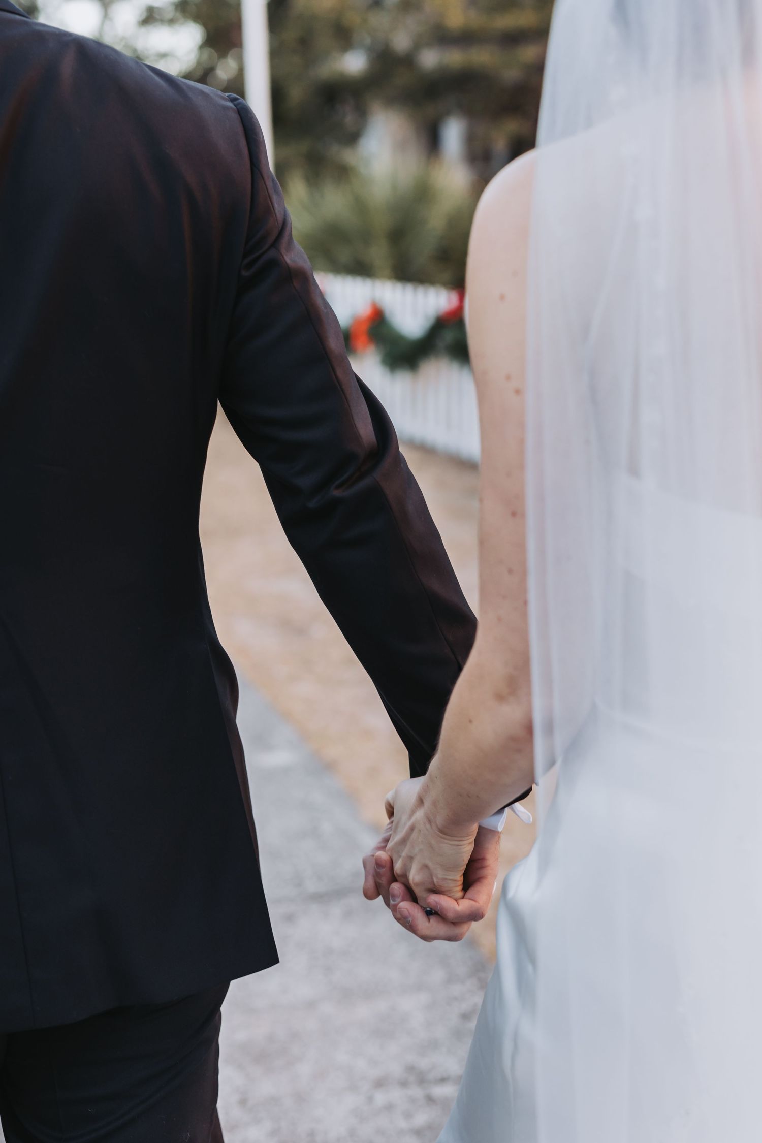 Close-up of bride and groom holding hands outdoors after wedding ceremony with holiday greenery backdrop.