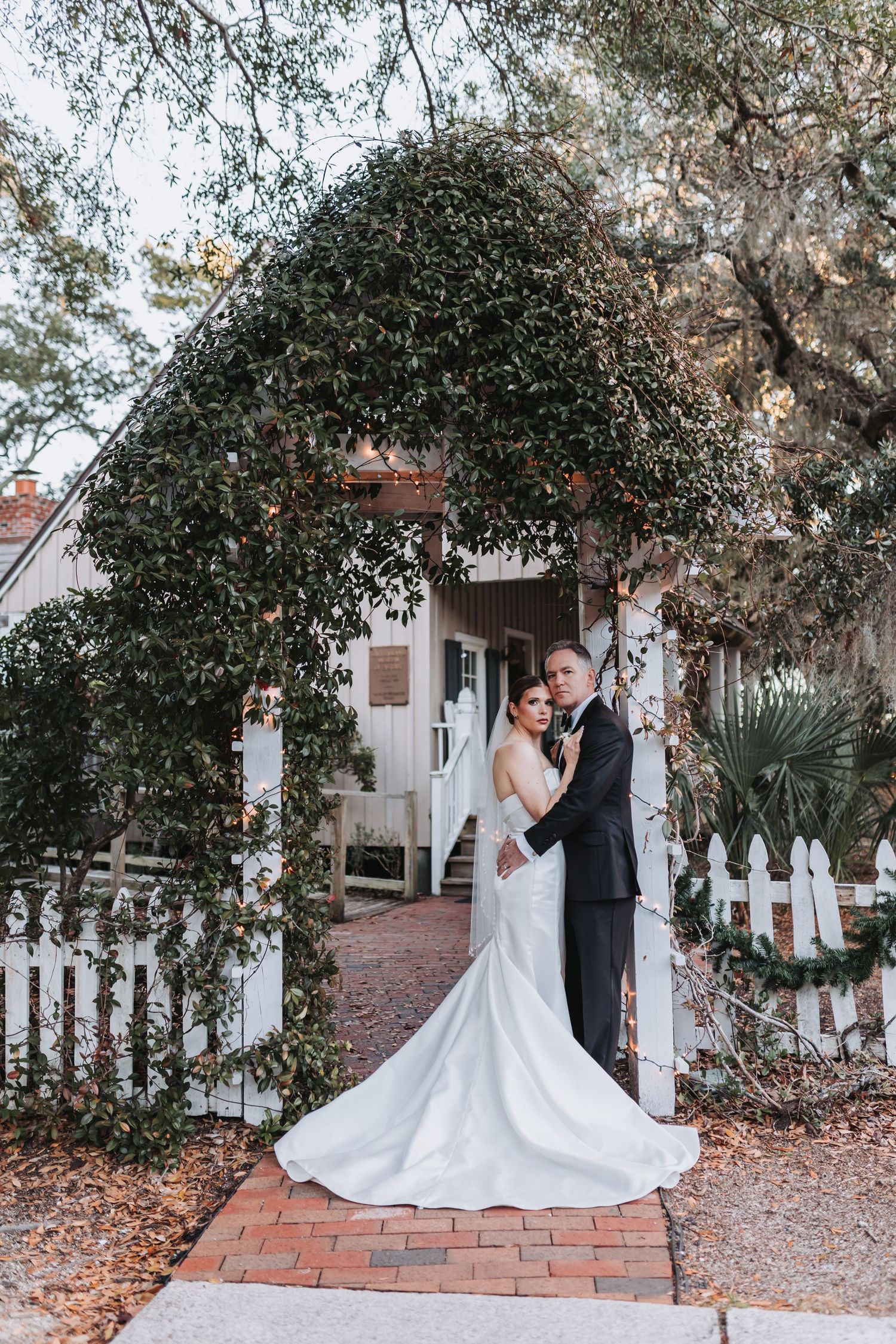 Bride and groom embracing under ivy-covered archway at white picket fence entrance at dusk.