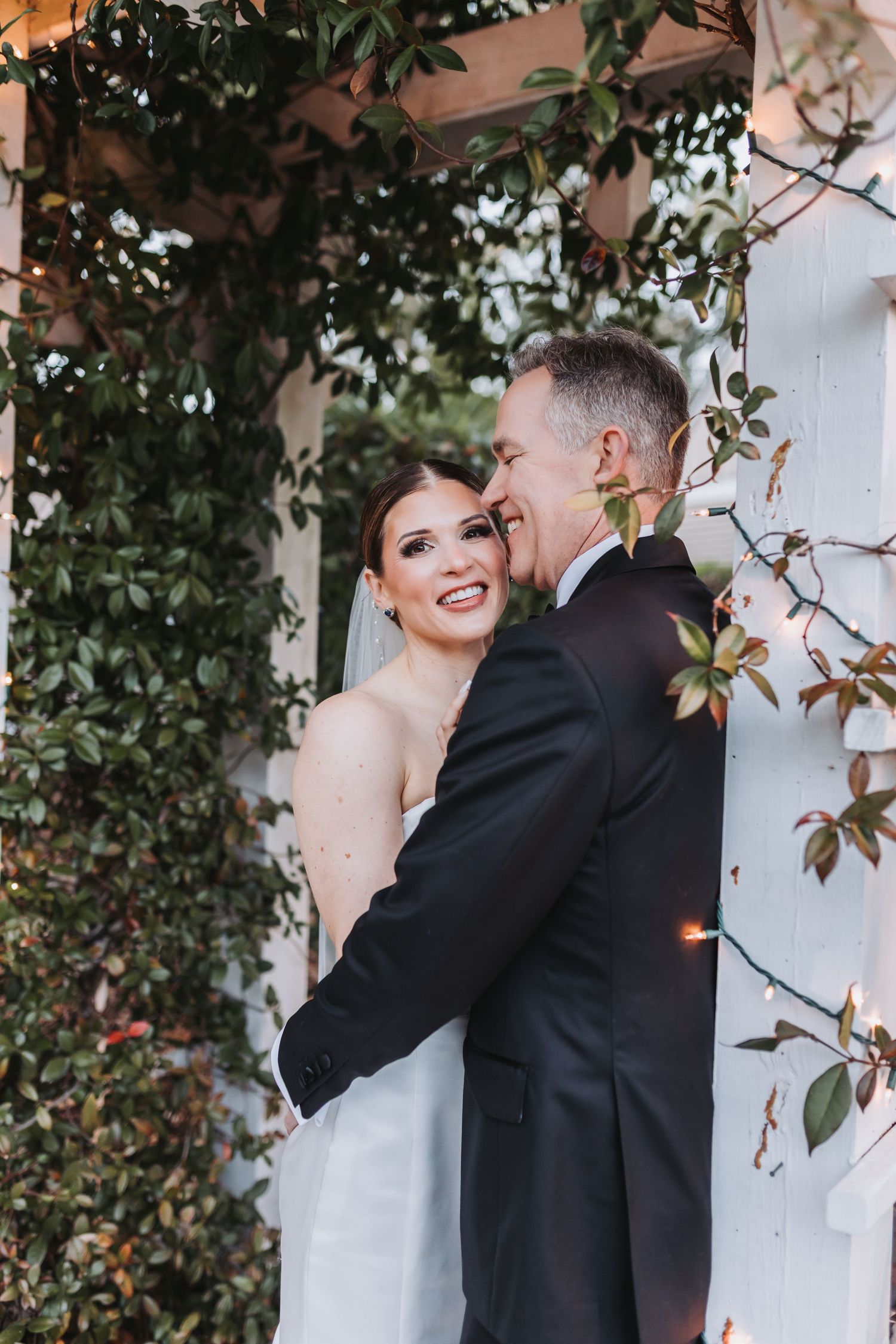 Groom embracing bride from behind as she smiles joyfully beneath a vine-covered white arbor with lights.