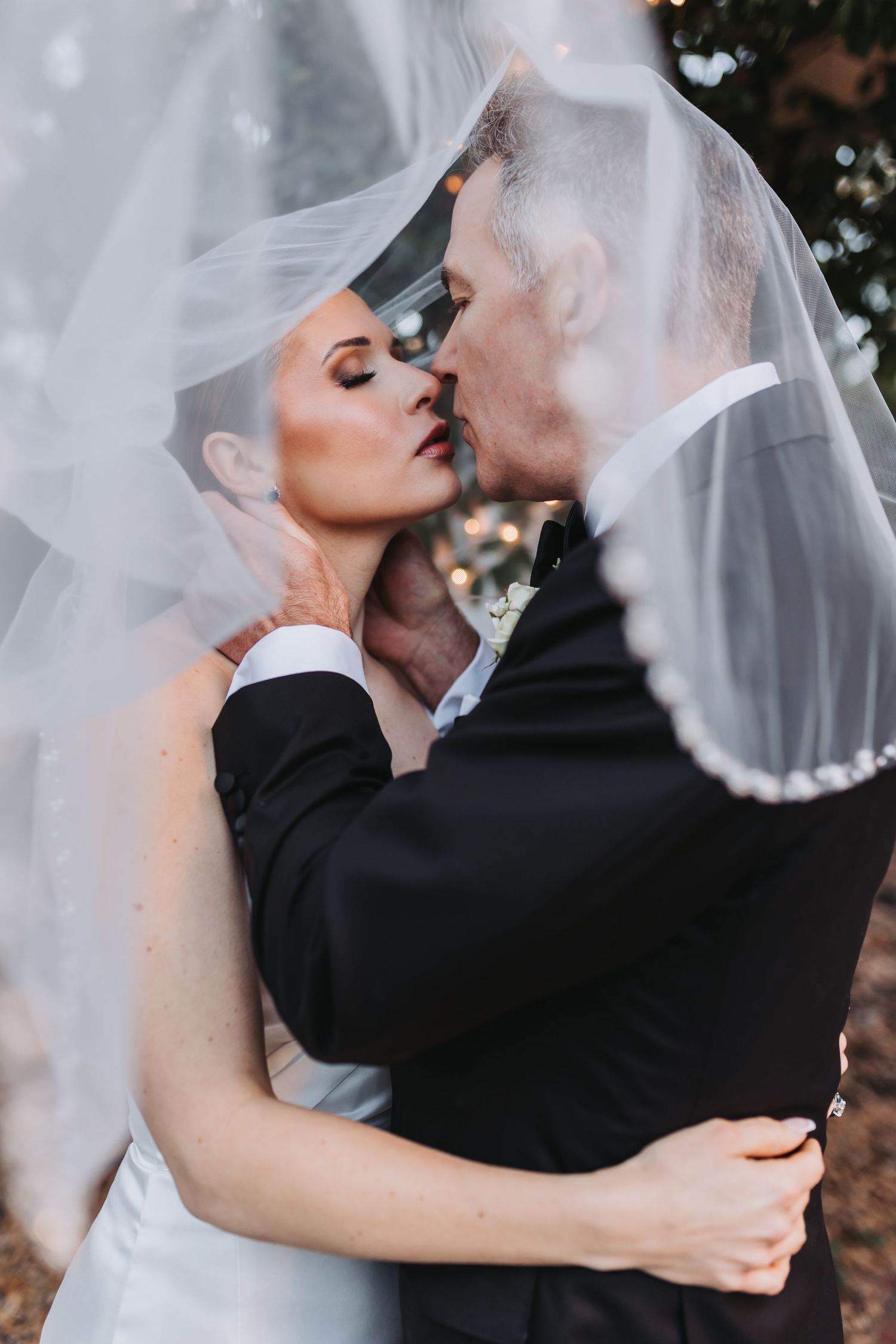 A bride and groom share an intimate kiss beneath a flowing white veil outdoors at golden hour.