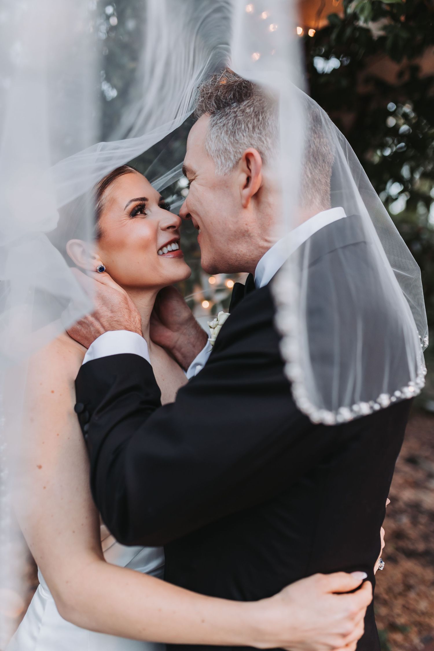 Romantic portrait of bride and groom sharing intimate moment beneath flowing wedding veil outdoors.