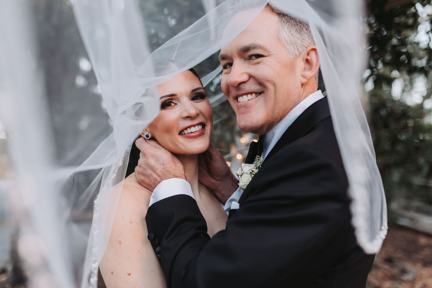 A bride and groom smile warmly under a flowing white veil on their wedding day.