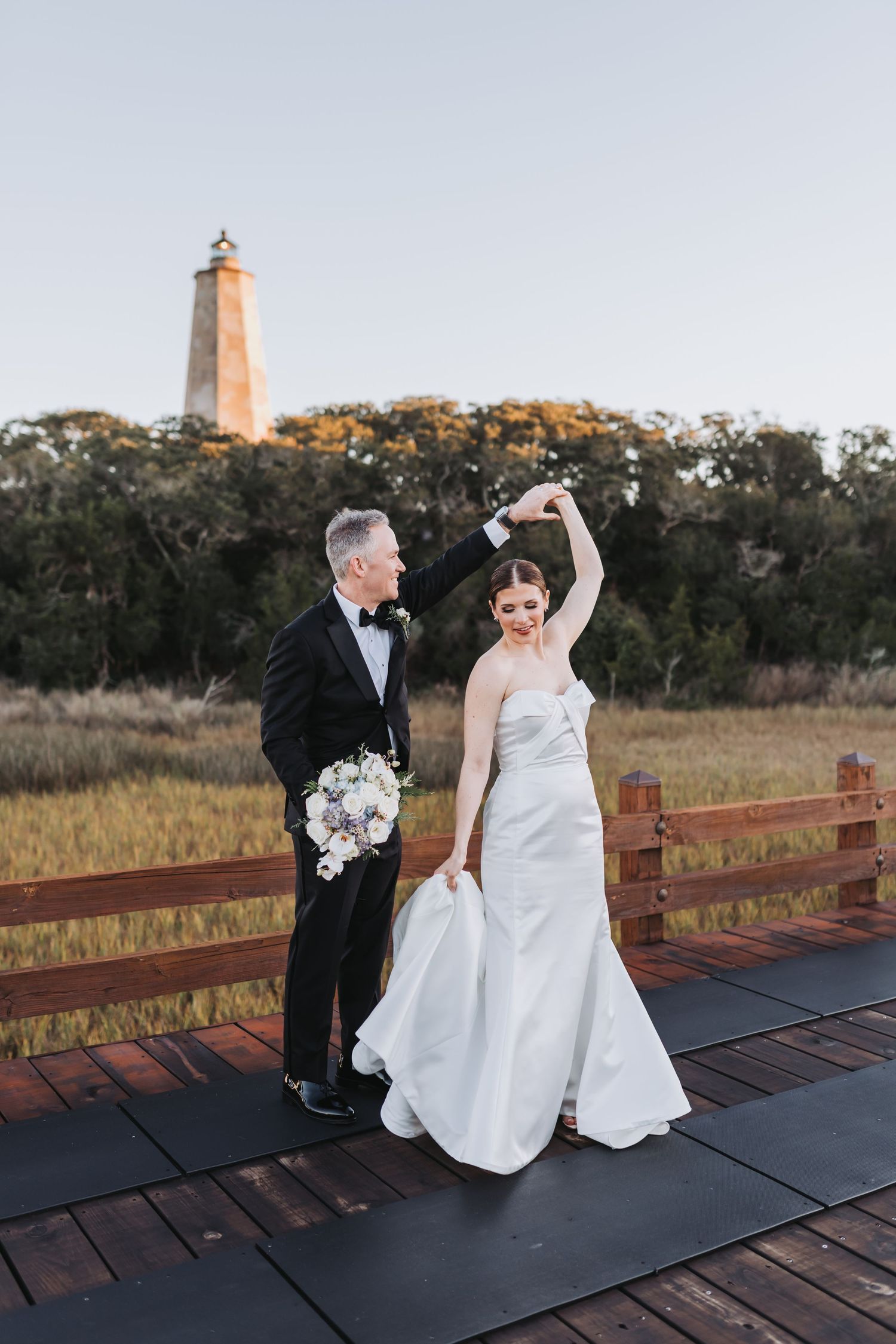 Groom twirls bride in elegant white dress on wooden deck with historic lighthouse in background.