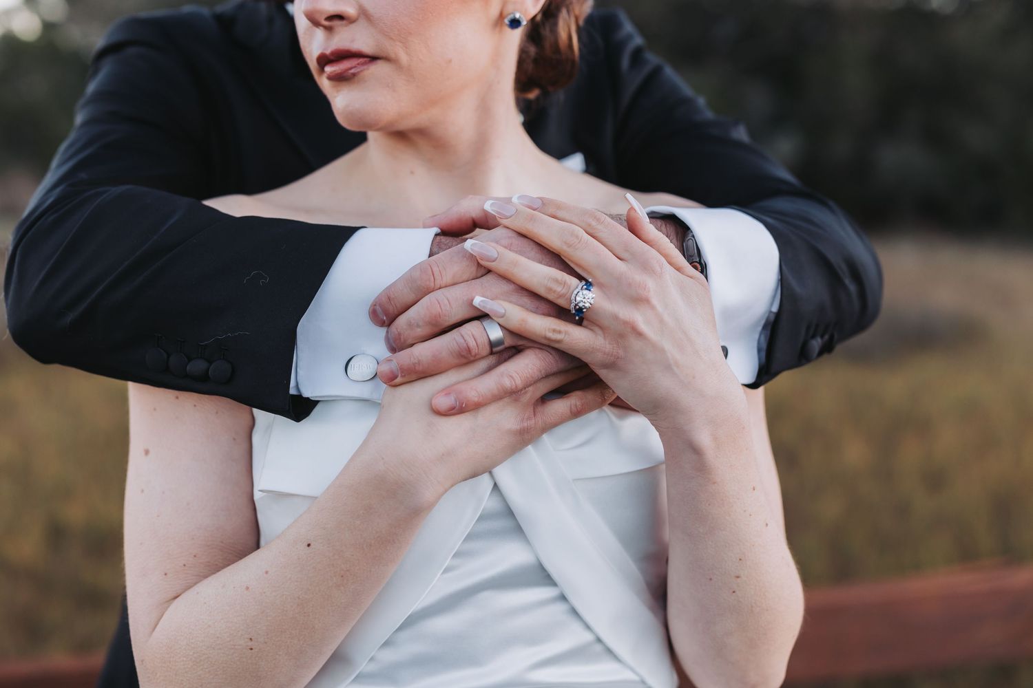 Close-up of groom embracing bride from behind showing wedding rings on her hands.