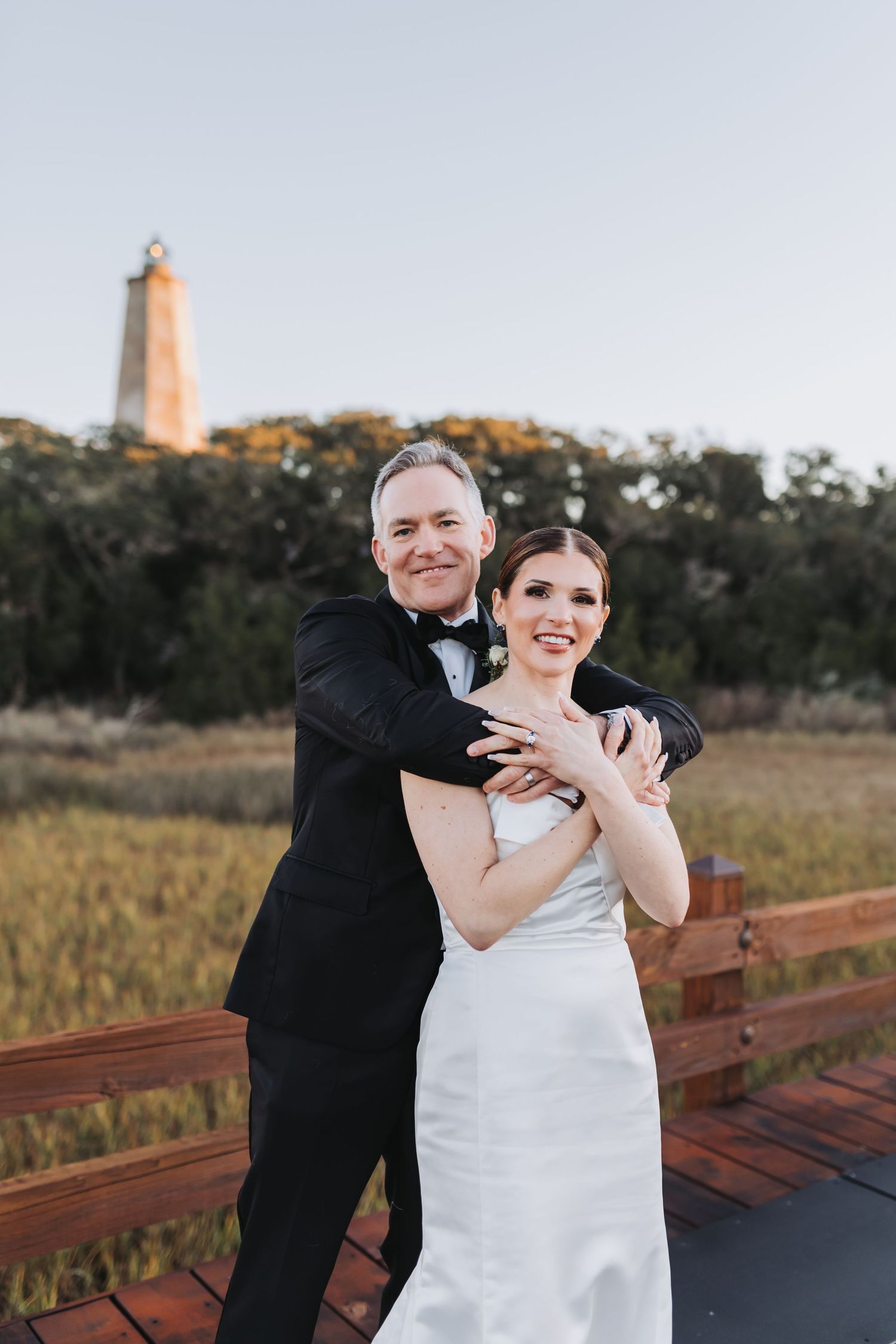 Bride and groom embracing on wooden deck at sunset with historic lighthouse in background at outdoor wedding.