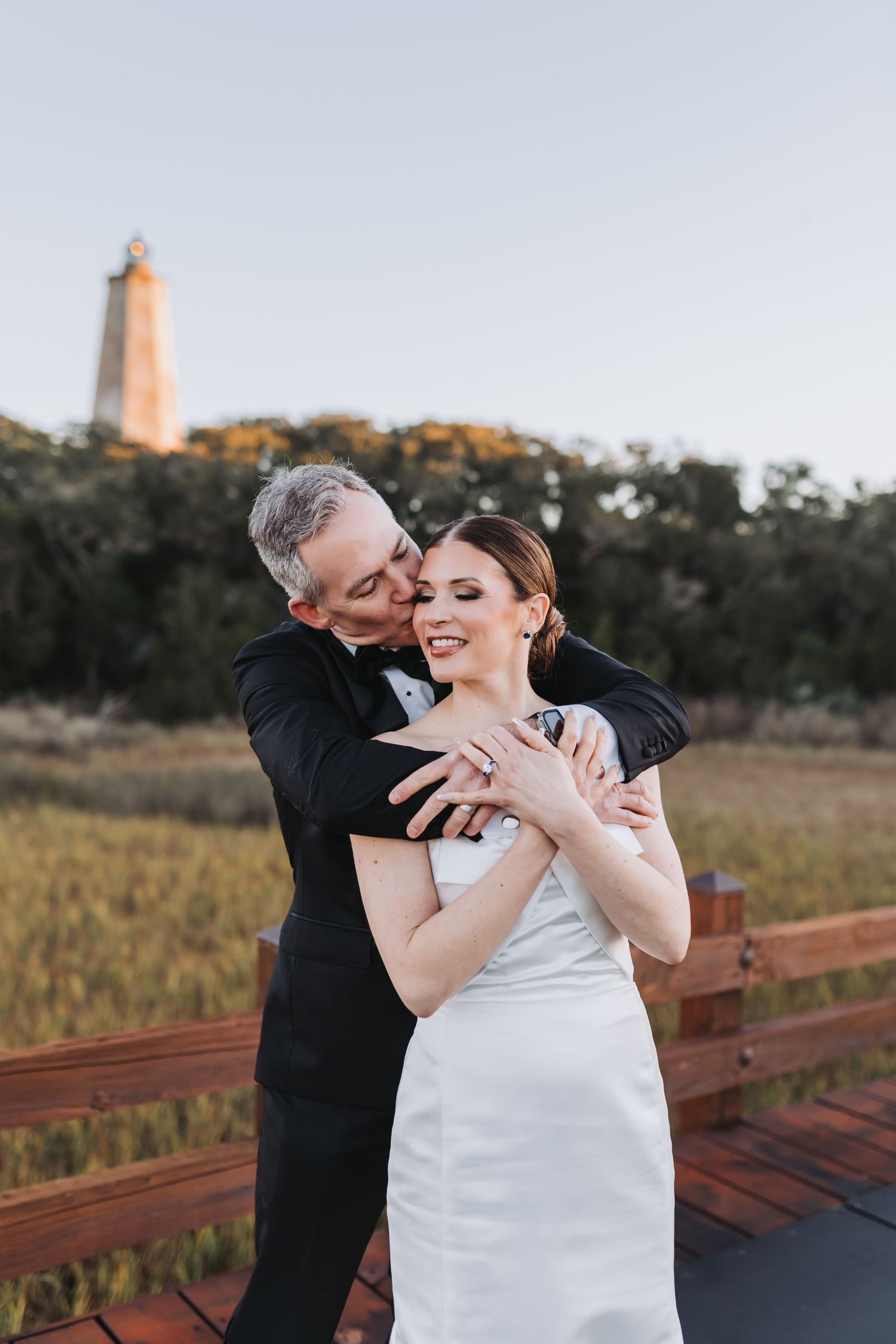 A groom in a black tuxedo kisses his bride in a white dress on a wooden deck near a lighthouse at sunset.