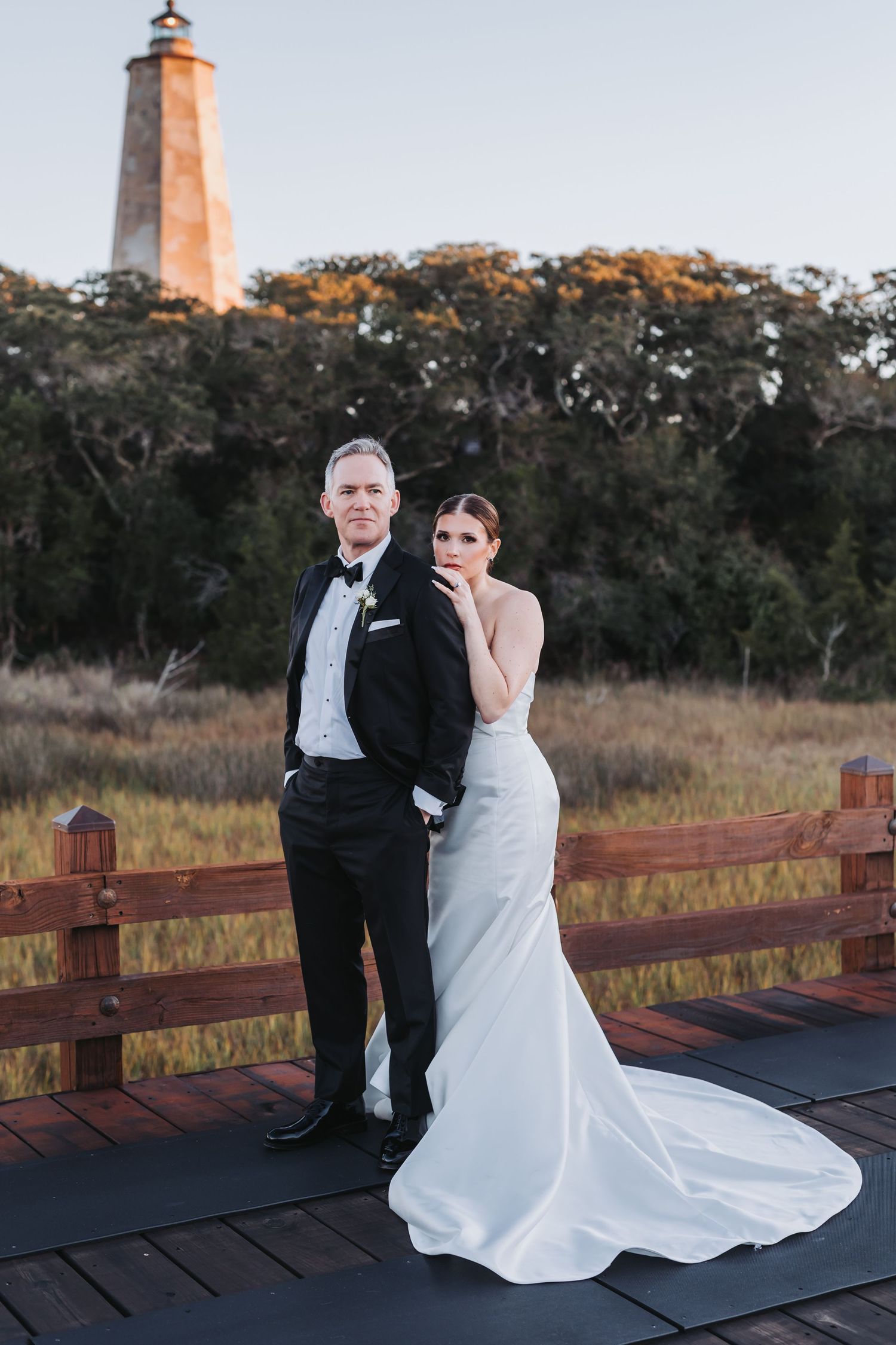 Elegant couple poses on wooden boardwalk with historic lighthouse and golden marshland at sunset.