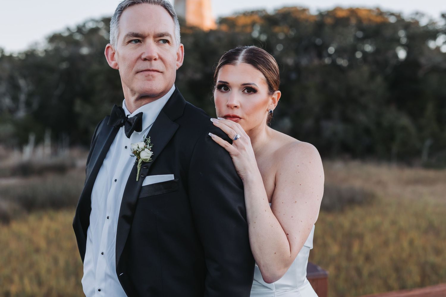 A bride leans against her groom in a tuxedo as they pose back-to-back in a golden marsh setting.