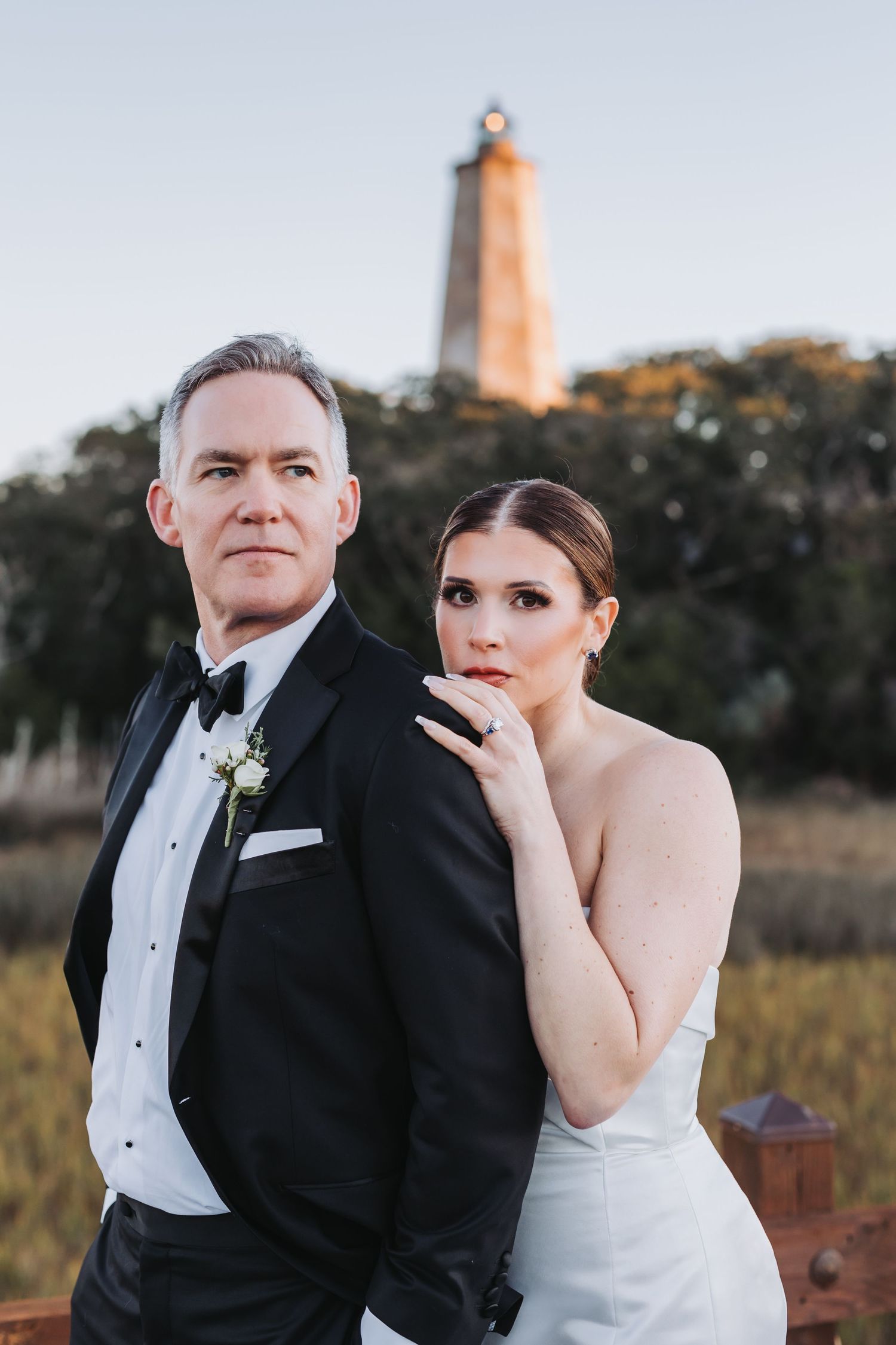 Bride and groom pose before historic lighthouse at golden hour in formal wedding attire.