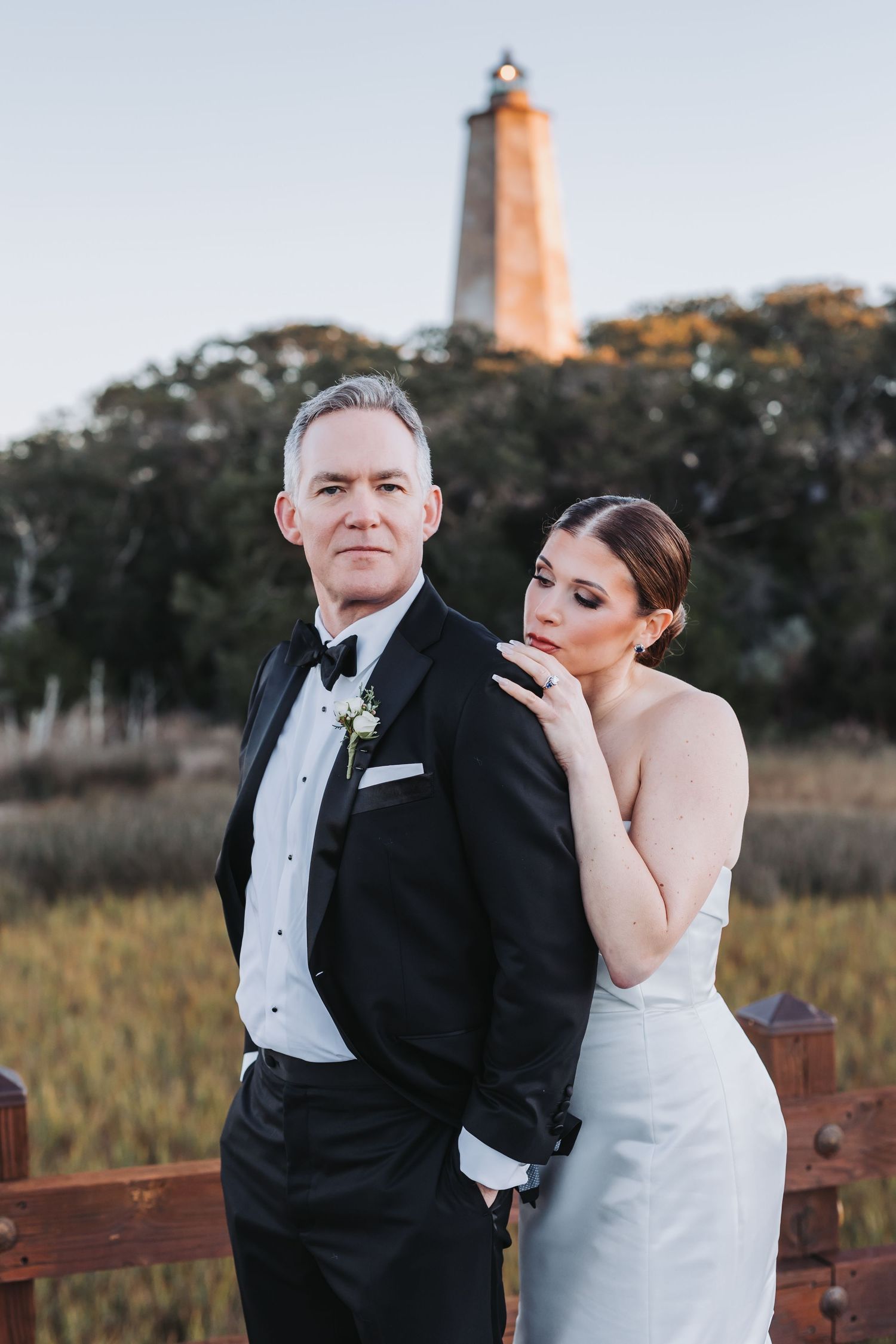 Elegant bride leans on groom in tuxedo with a historic lighthouse backdrop at their coastal wedding venue.